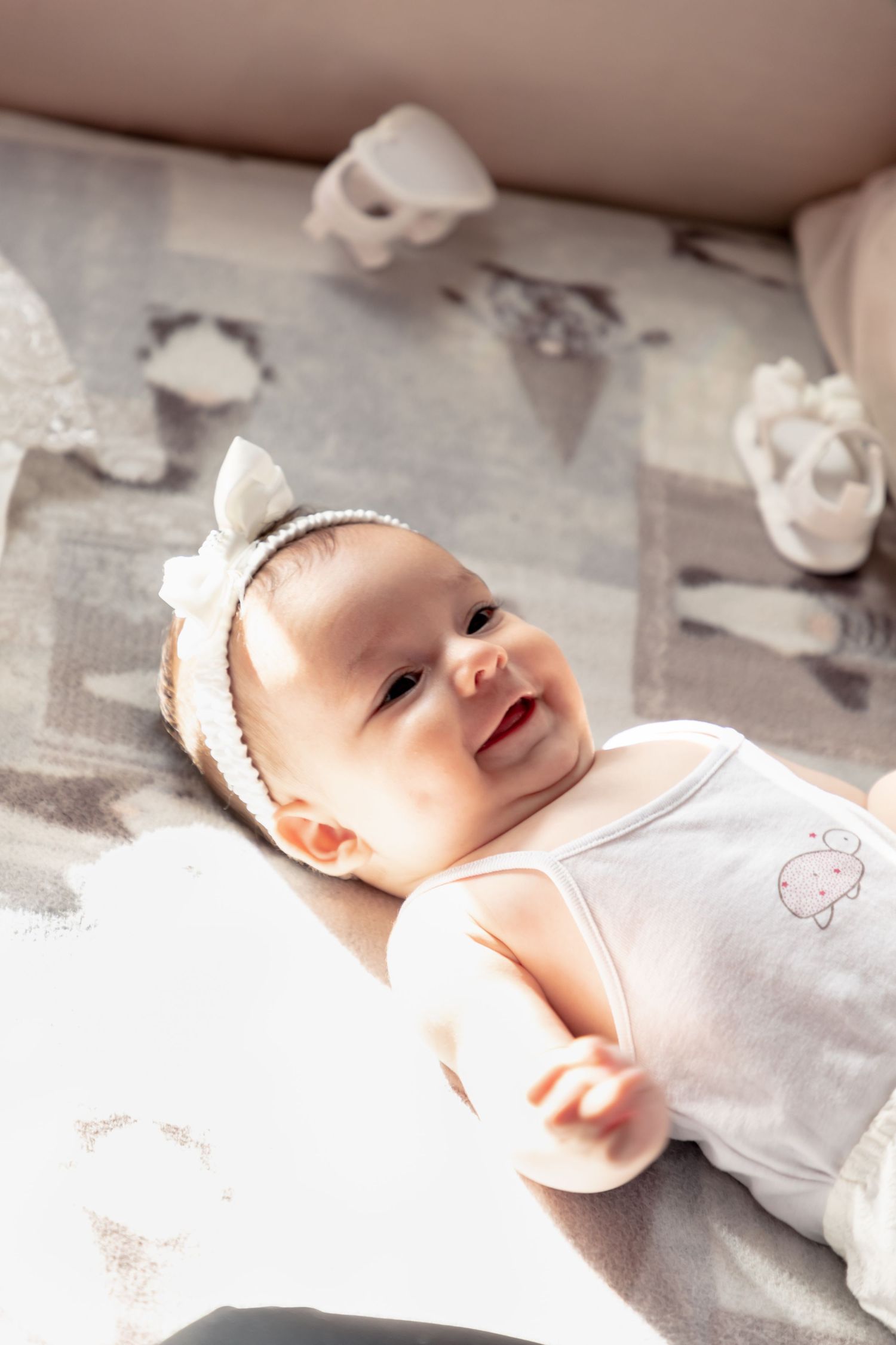 Smiling baby girl wearing a white headband bow lying on a soft grey patterned blanket.
