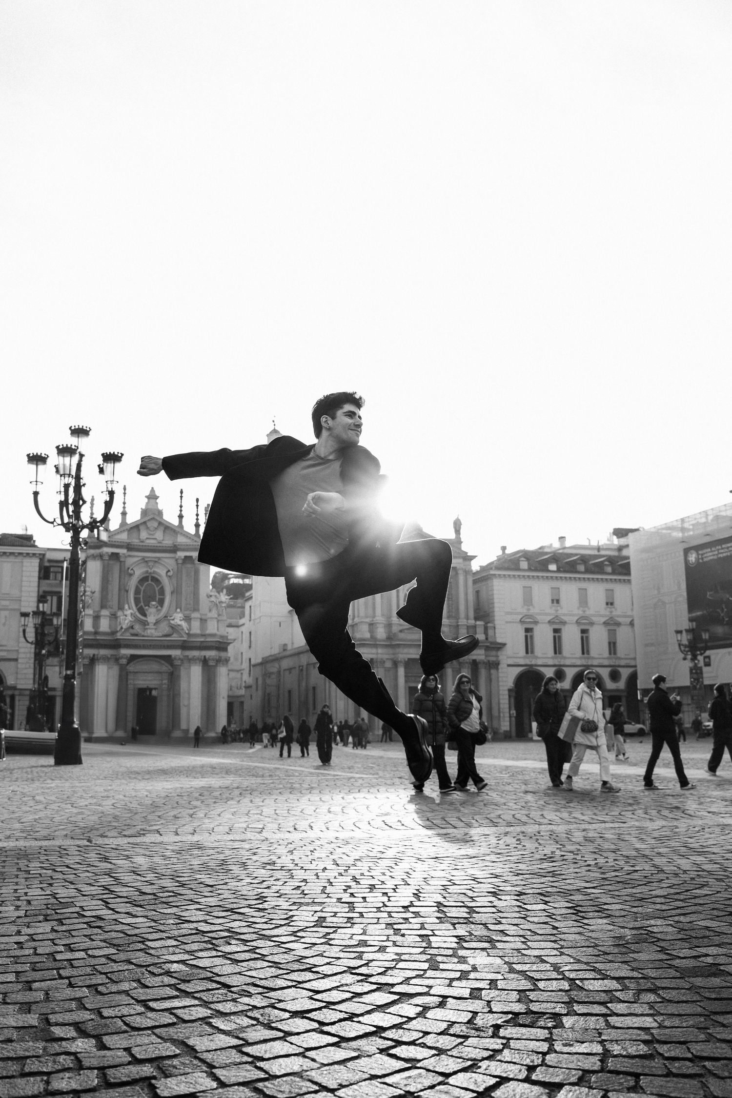 Black and white shot of male dancer leaping in sunlit historic European city square.
