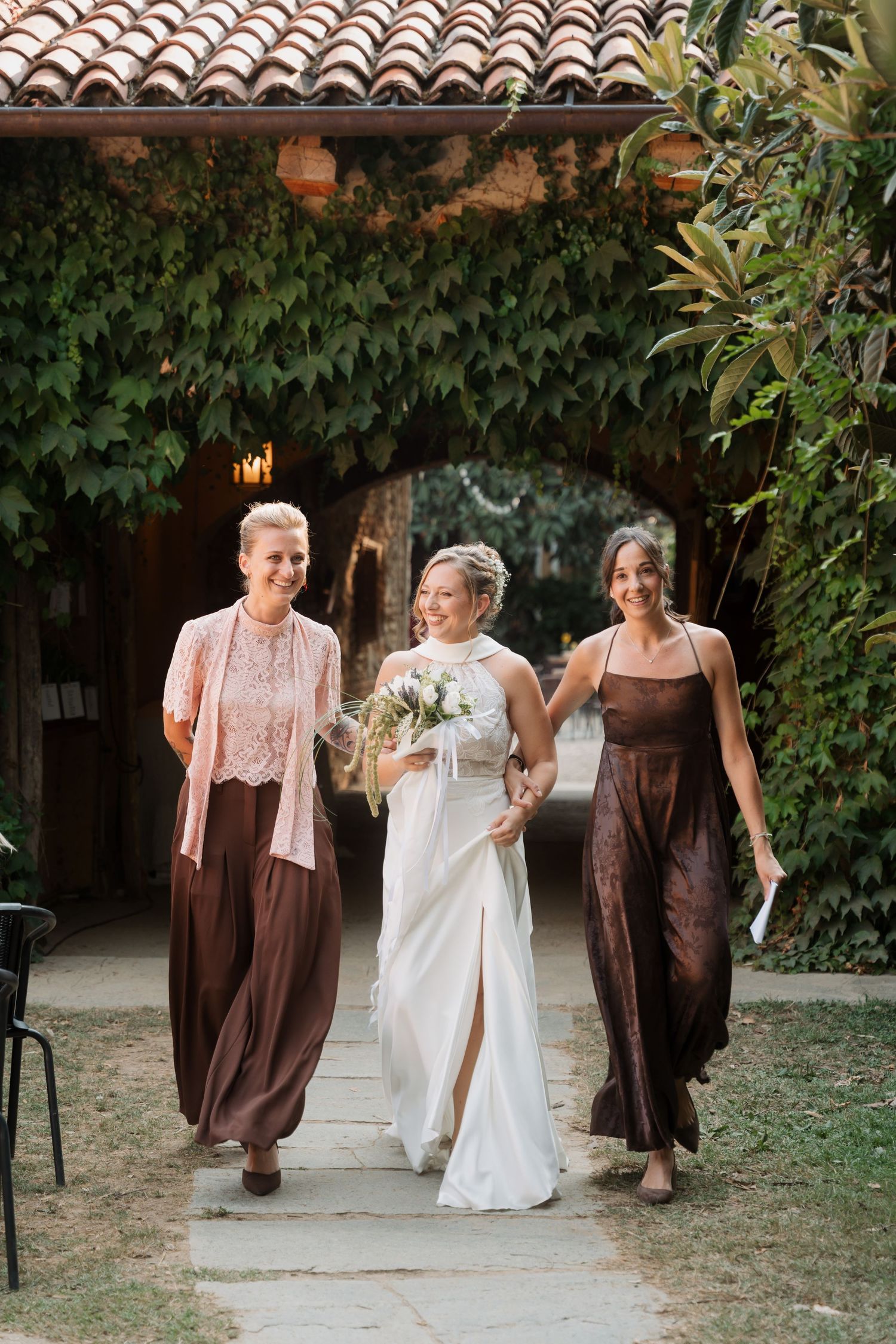 Bride in white gown walks with two bridesmaids in brown dresses through ivy-covered archway at outdoor wedding venue.