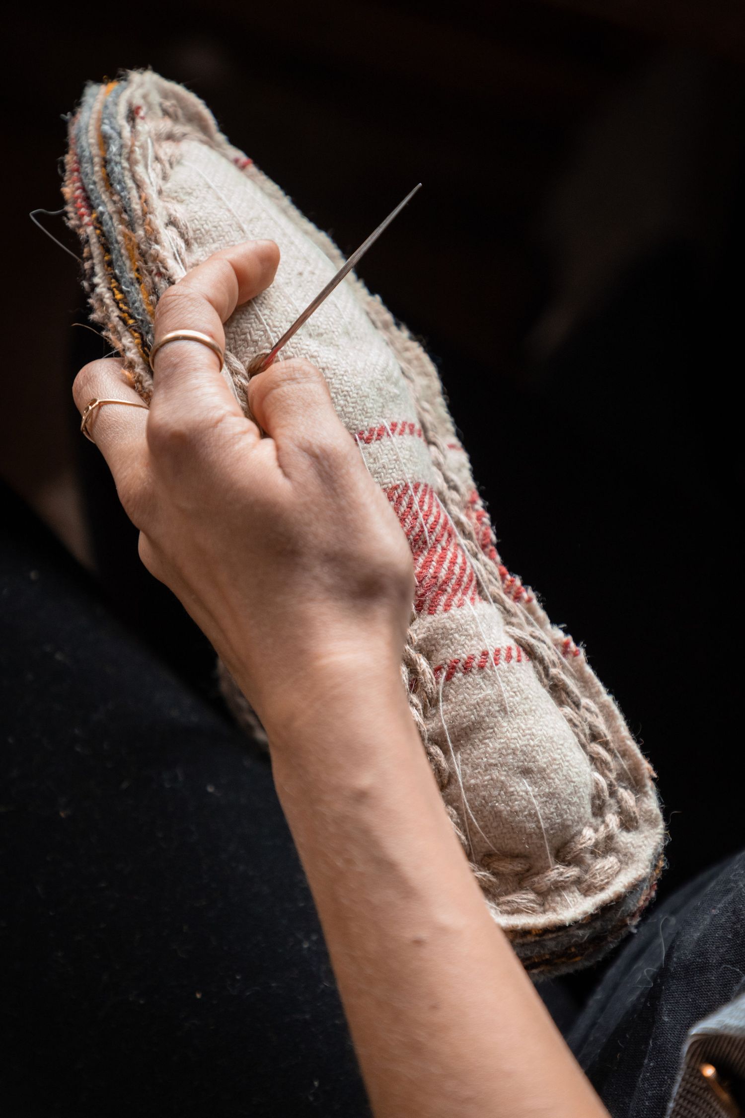 Close-up of hands sewing a traditional espadrille shoe sole with a needle, wearing delicate gold rings.
