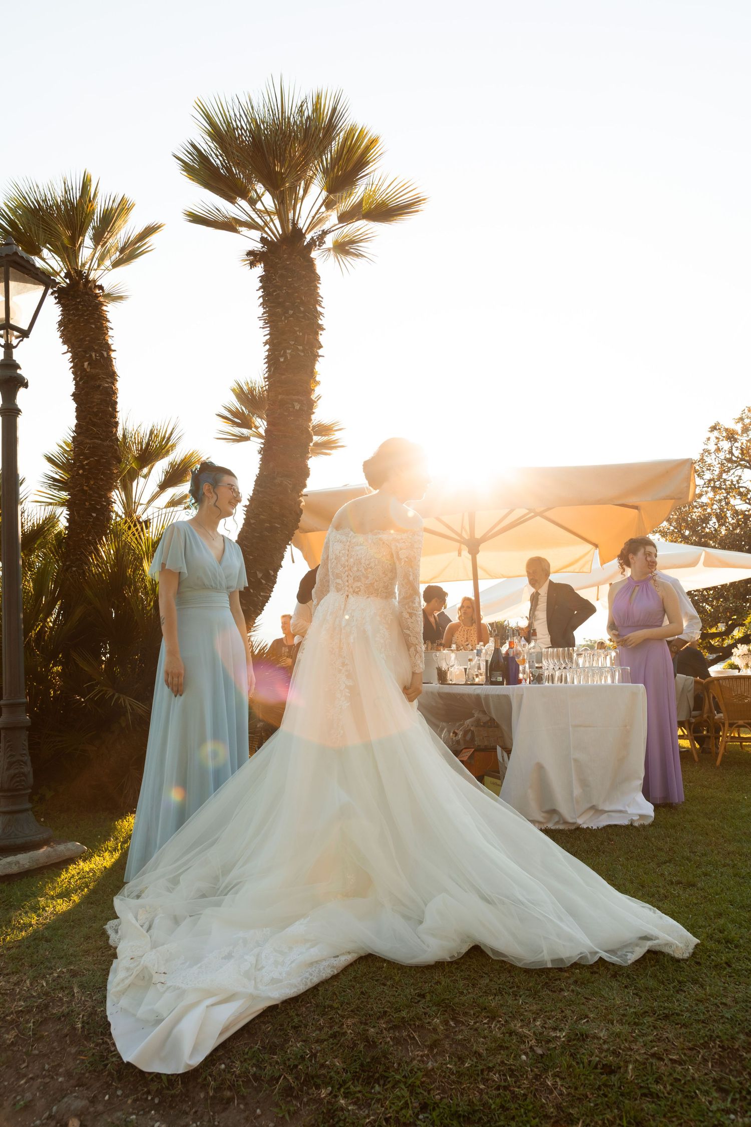 Bride in elegant lace gown with long train stands in garden at golden hour with wedding guests.