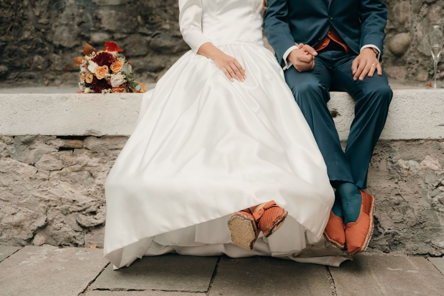 Close-up of bride in white gown and groom in blue suit with orange shoes seated on stone bench.