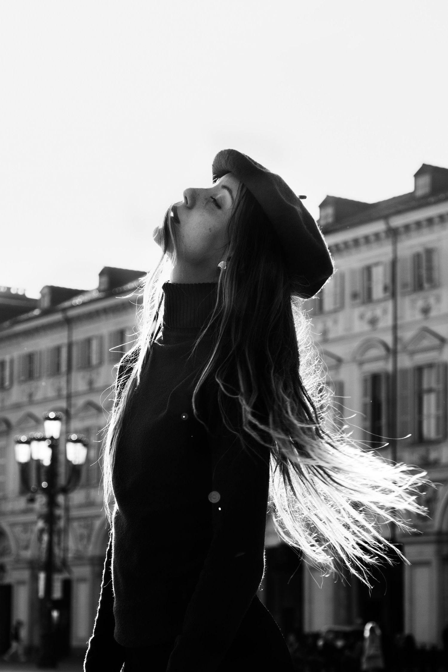 Woman with long flowing hair tilting head back in sunlight before classic European building, black and white.
