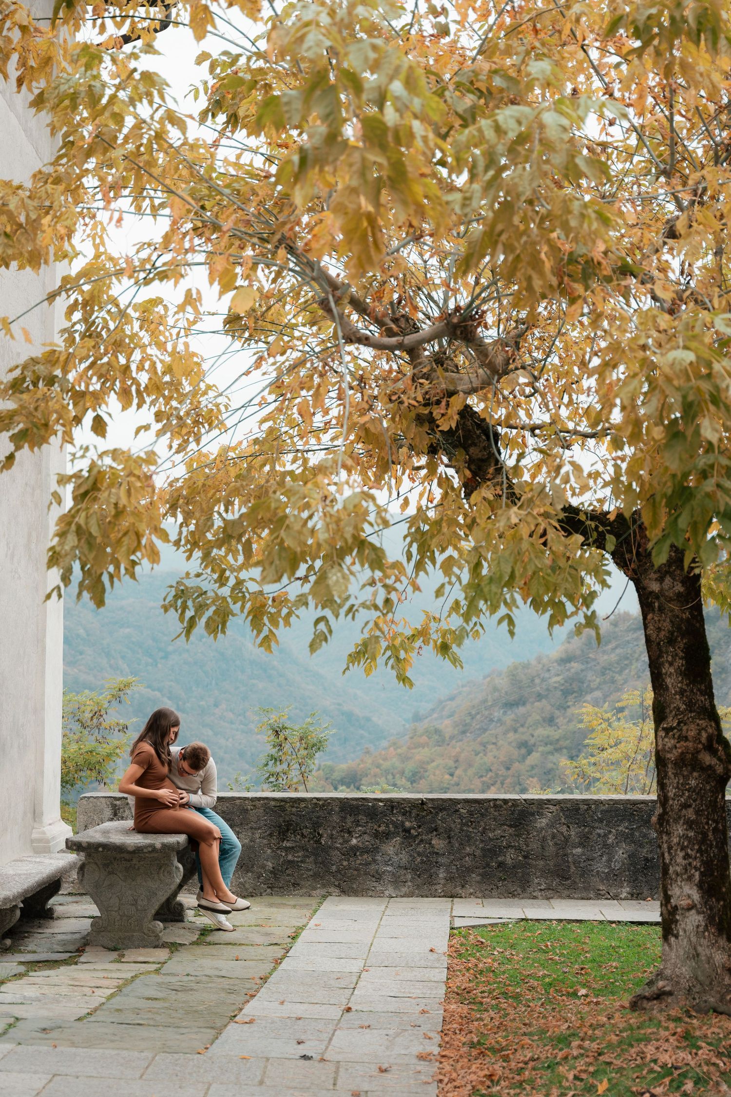 Mother and child sitting on a stone bench under golden autumn tree with mountain view.