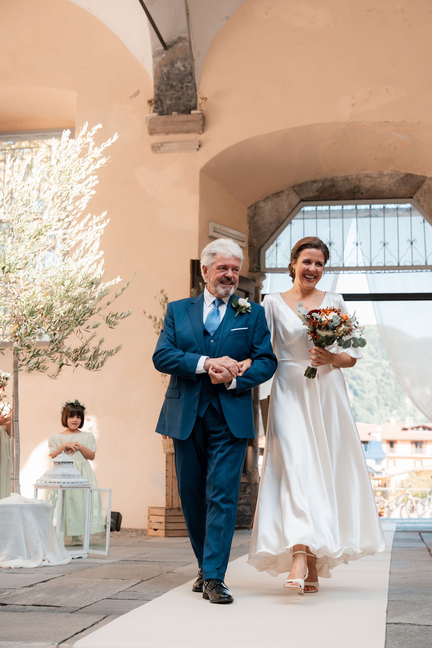 Father escorts smiling bride in white gown down outdoor aisle holding colorful bridal bouquet.