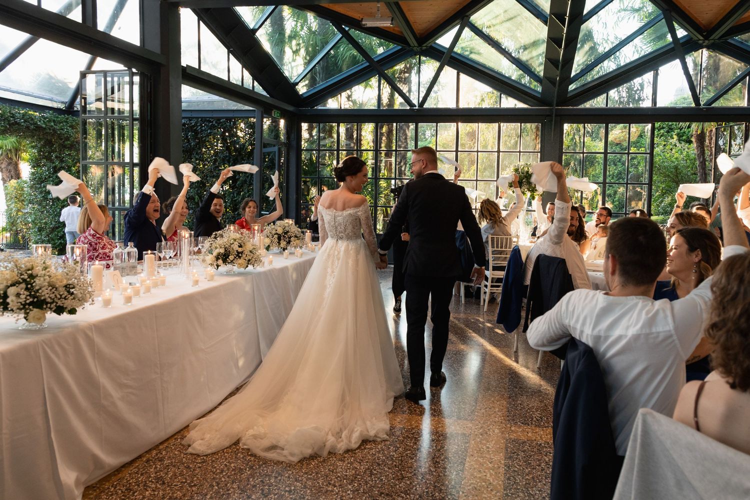Bride and groom enter wedding reception in a glass greenhouse venue as guests celebrate with raised hands.