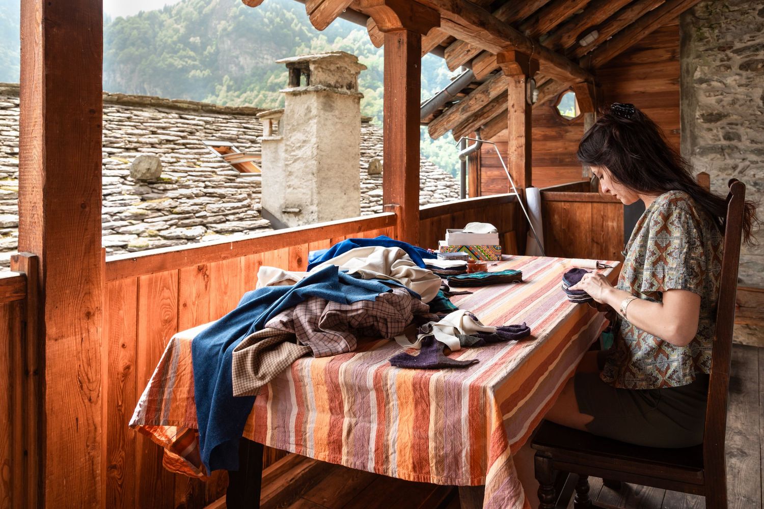 A woman works at a wooden table on a rustic balcony overlooking a stone village.