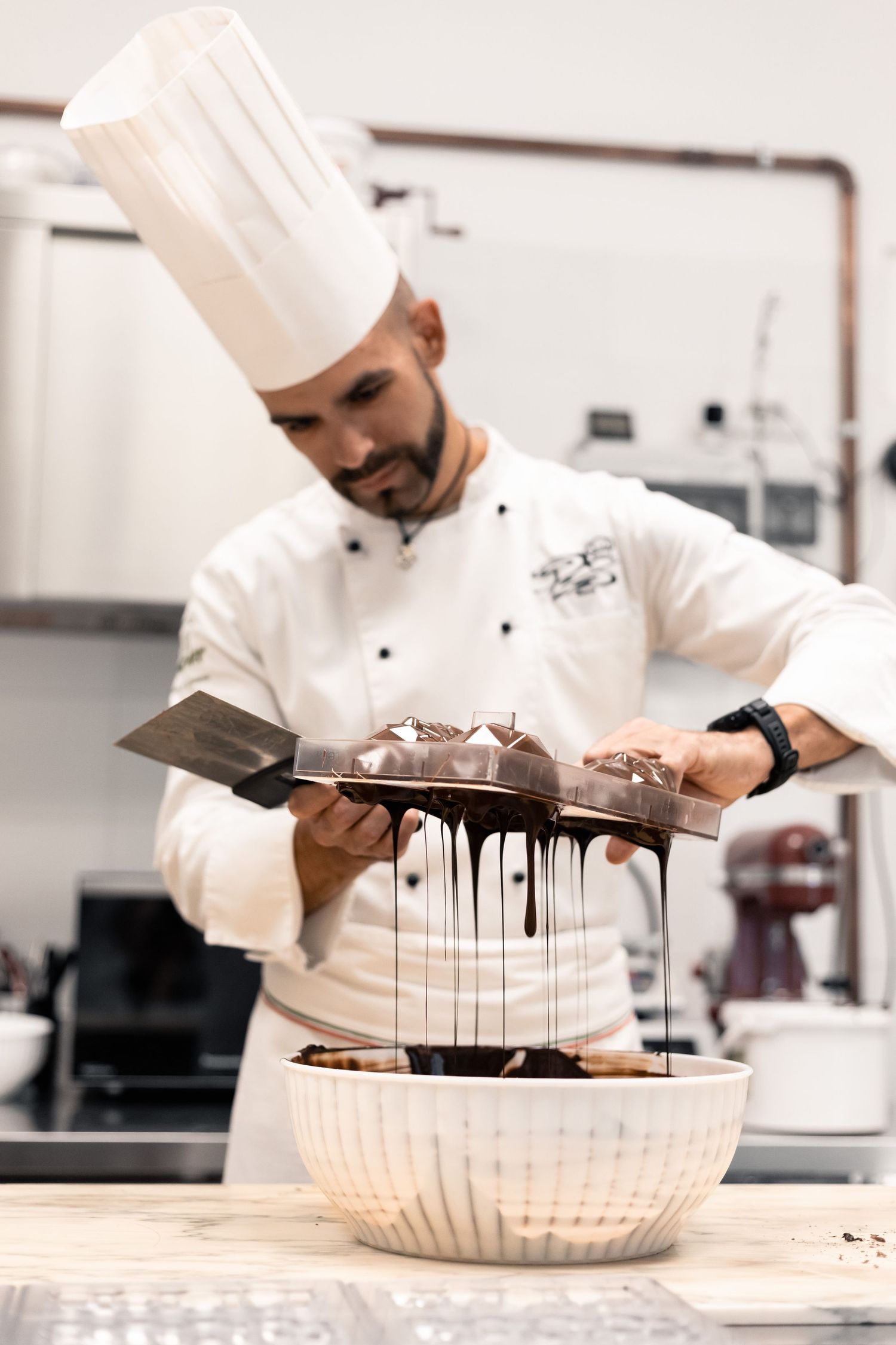Professional pastry chef in white uniform drizzling chocolate ganache from metal scraper into bowl.