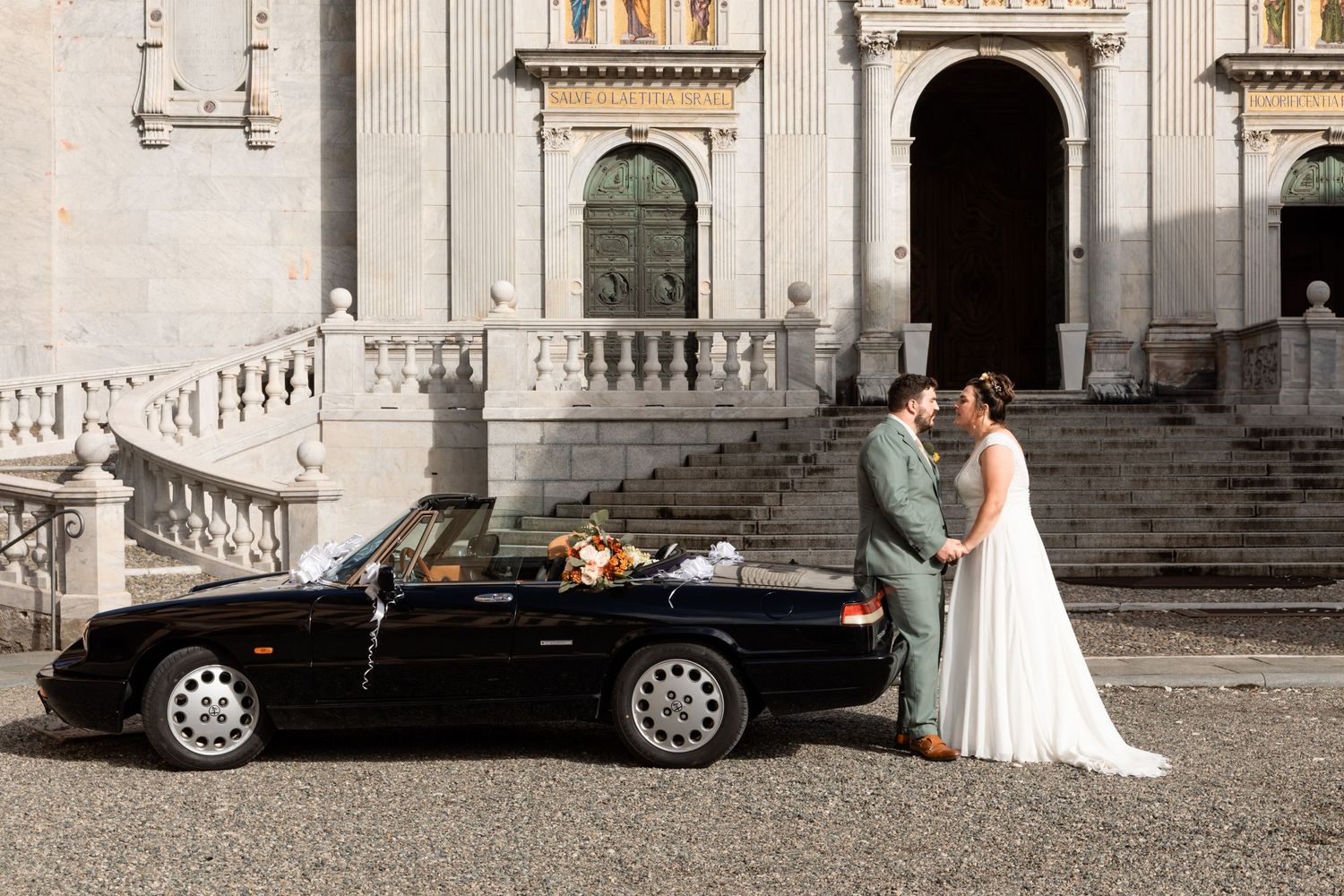 Bride and groom posing beside a classic black convertible car in front of an elegant European church.