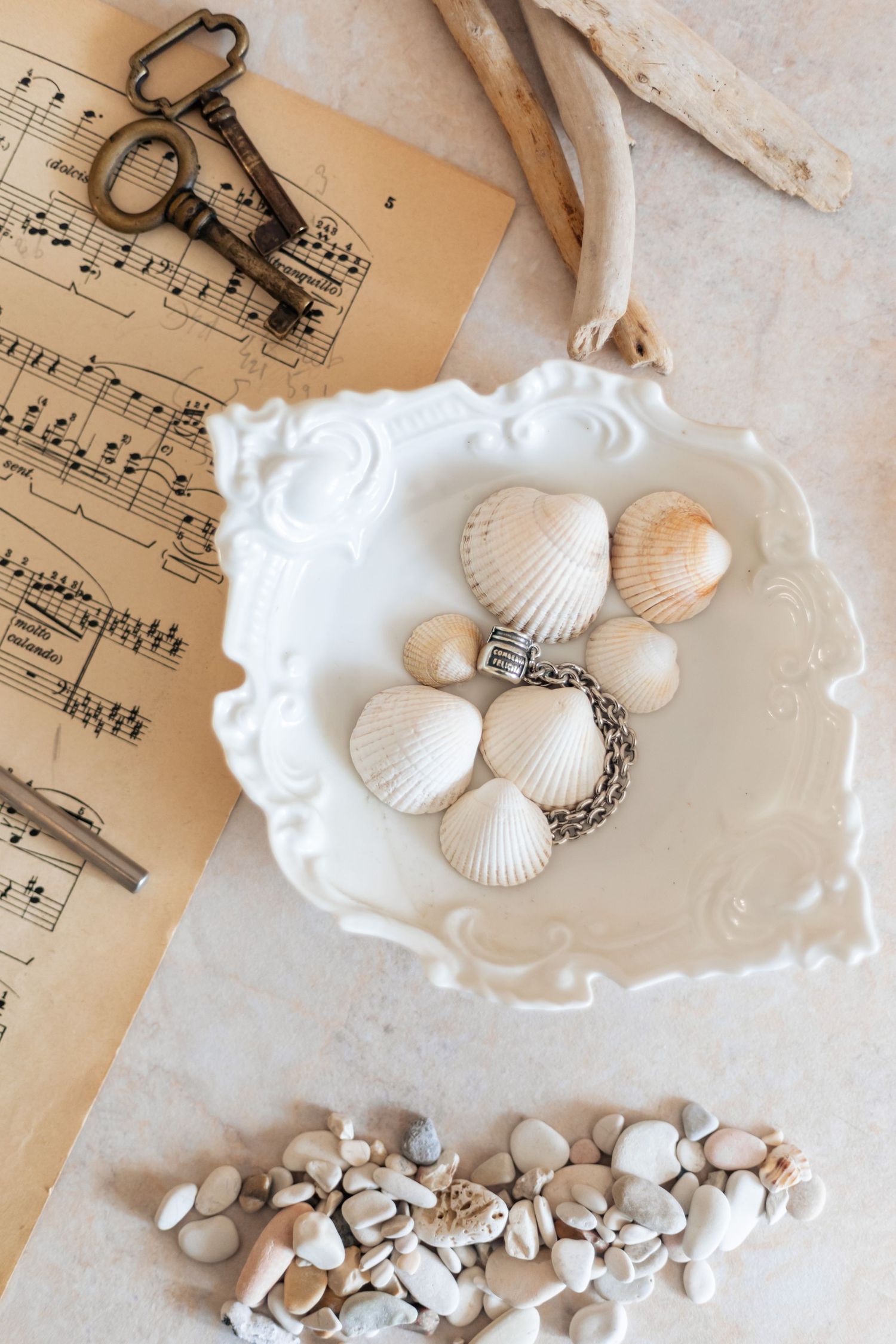 Vintage music sheets with antique keys, seashells on white ornate dish, and pebbles arranged on marble surface.