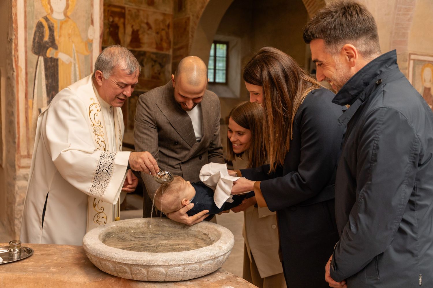 Catholic priest baptizing infant at stone font surrounded by family in historic frescoed church.
