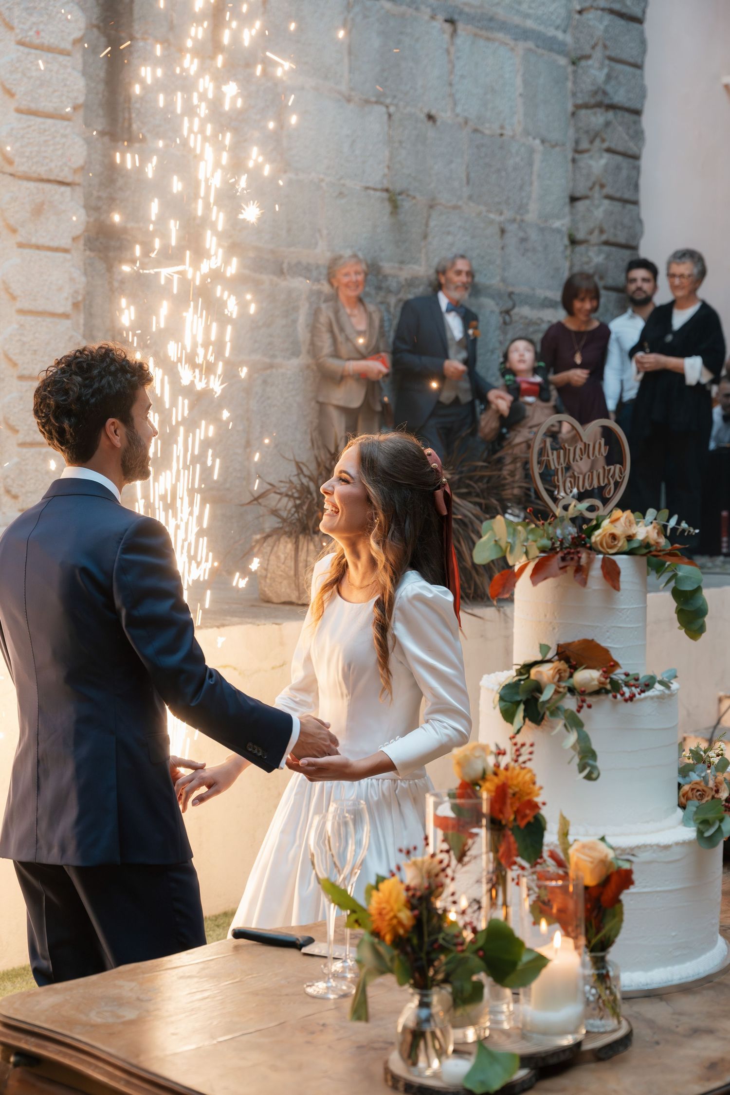 Bride and groom hold hands beside decorated wedding cake as sparklers illuminate elegant outdoor stone venue.