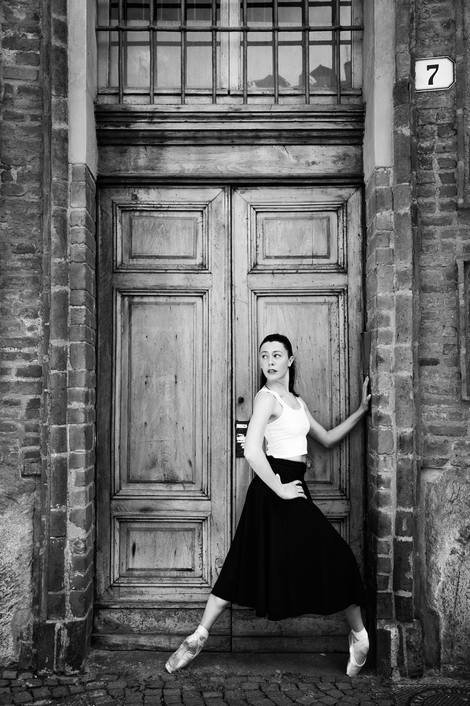 Elegant ballerina in black skirt and white top posing on pointe by old wooden door, black and white.