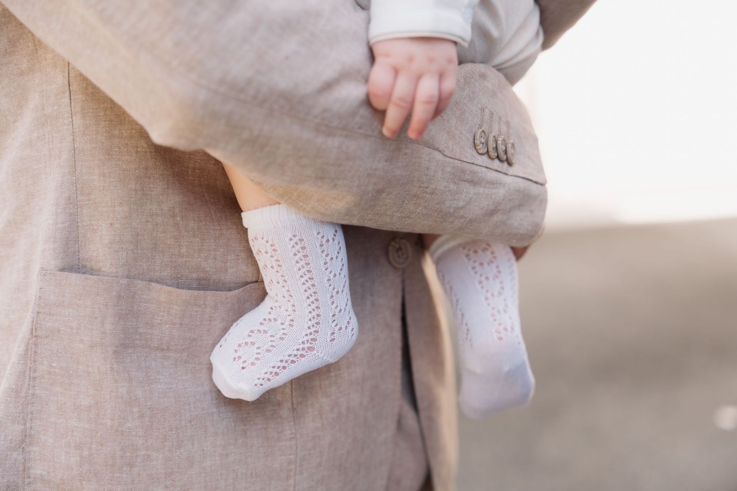 Close-up of a baby wearing delicate white lace socks being held by parent in beige linen outfit.