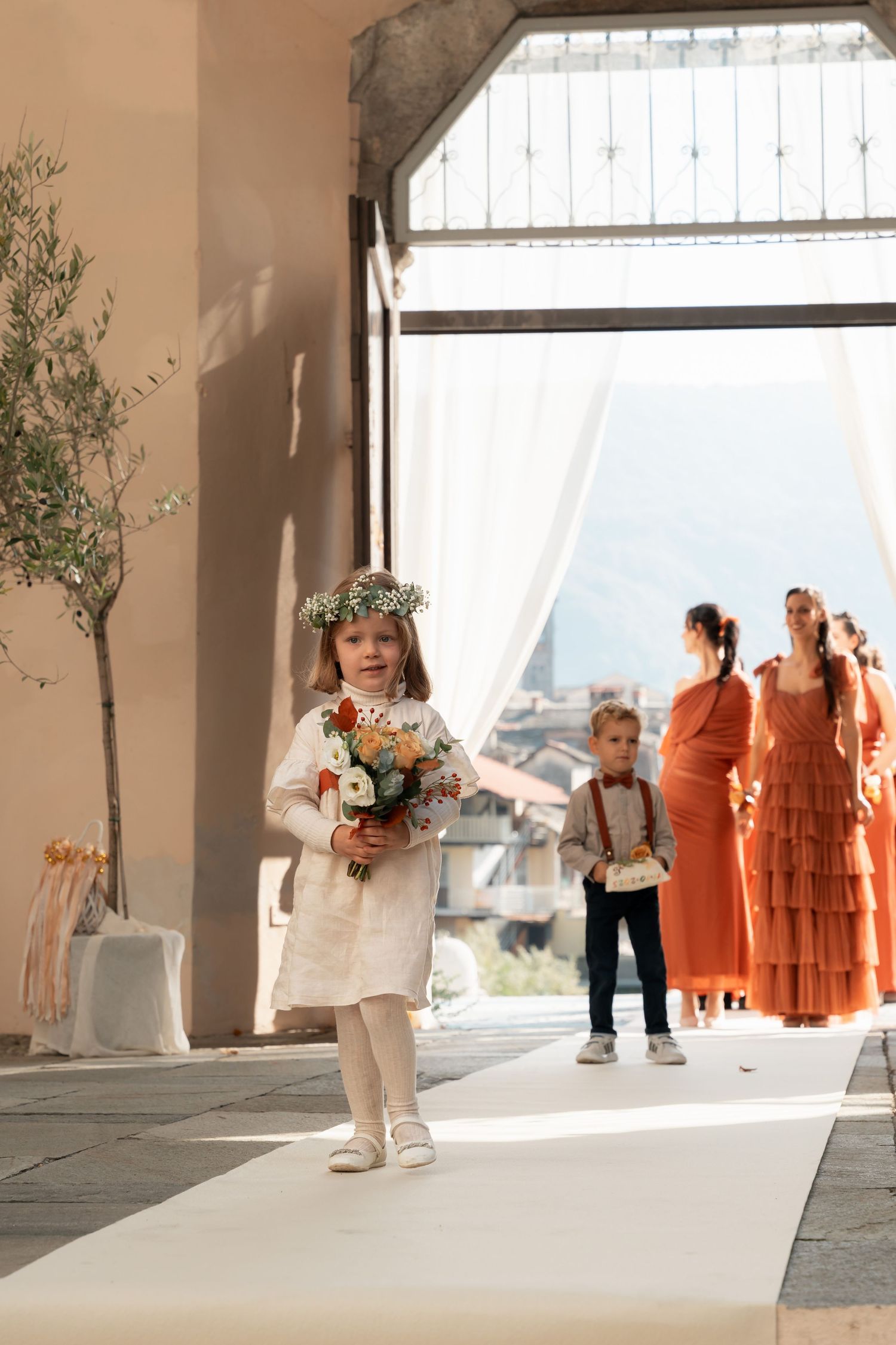 Young flower girl in white dress with floral crown walks down aisle holding bouquet at wedding ceremony.