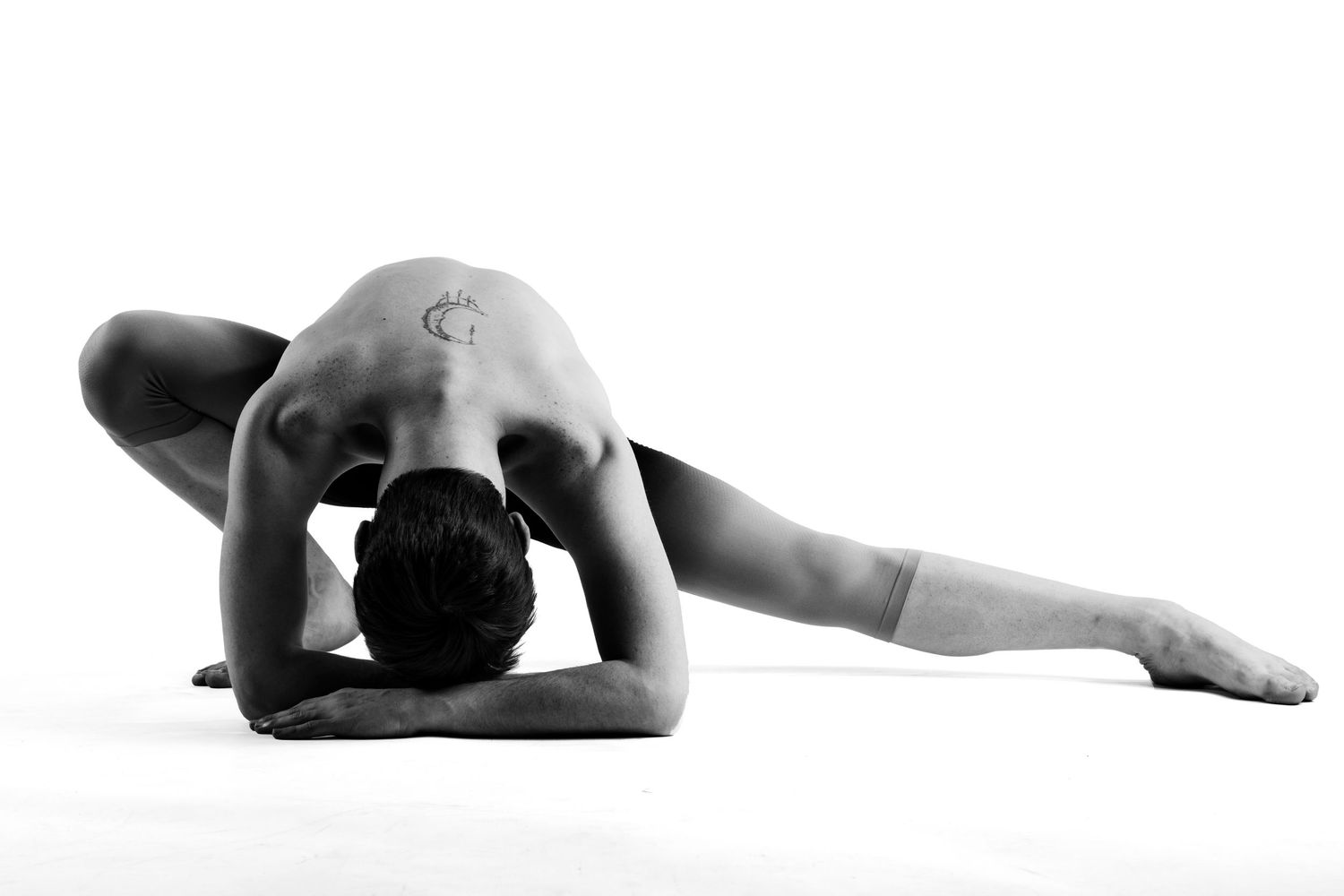 Male dancer in deep forward stretch pose with back arched low to floor in black and white.