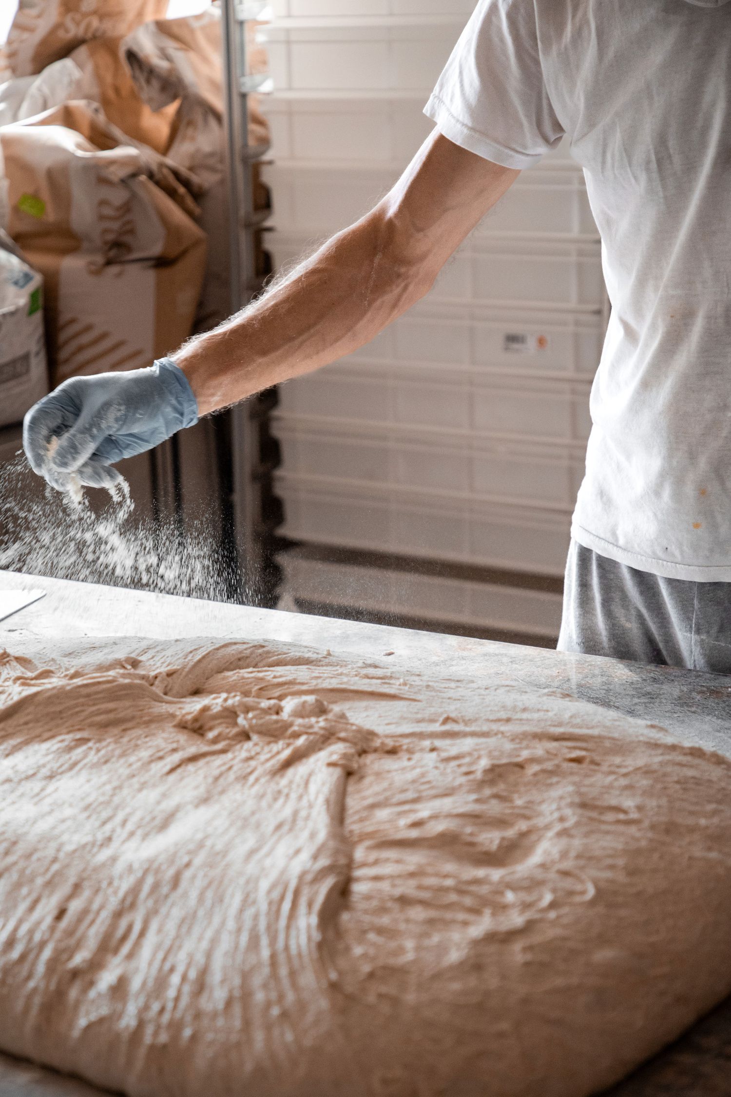 Baker in white t-shirt sprinkling flour over large batch of bread dough on work table.