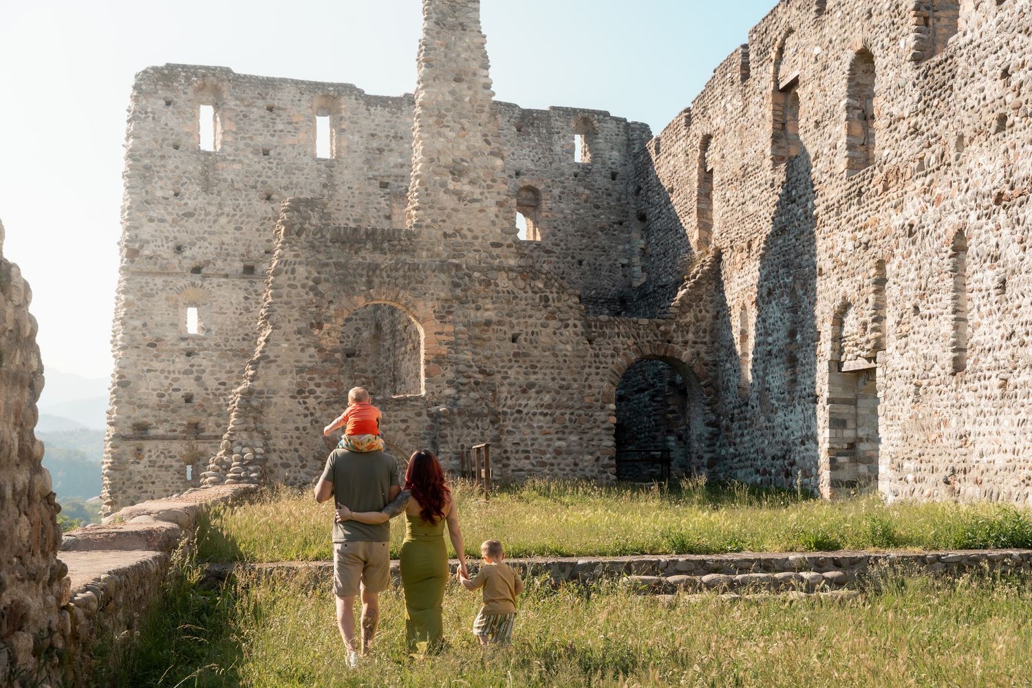 Family walking toward ancient stone castle ruins through tall green grass in warm sunlight.
