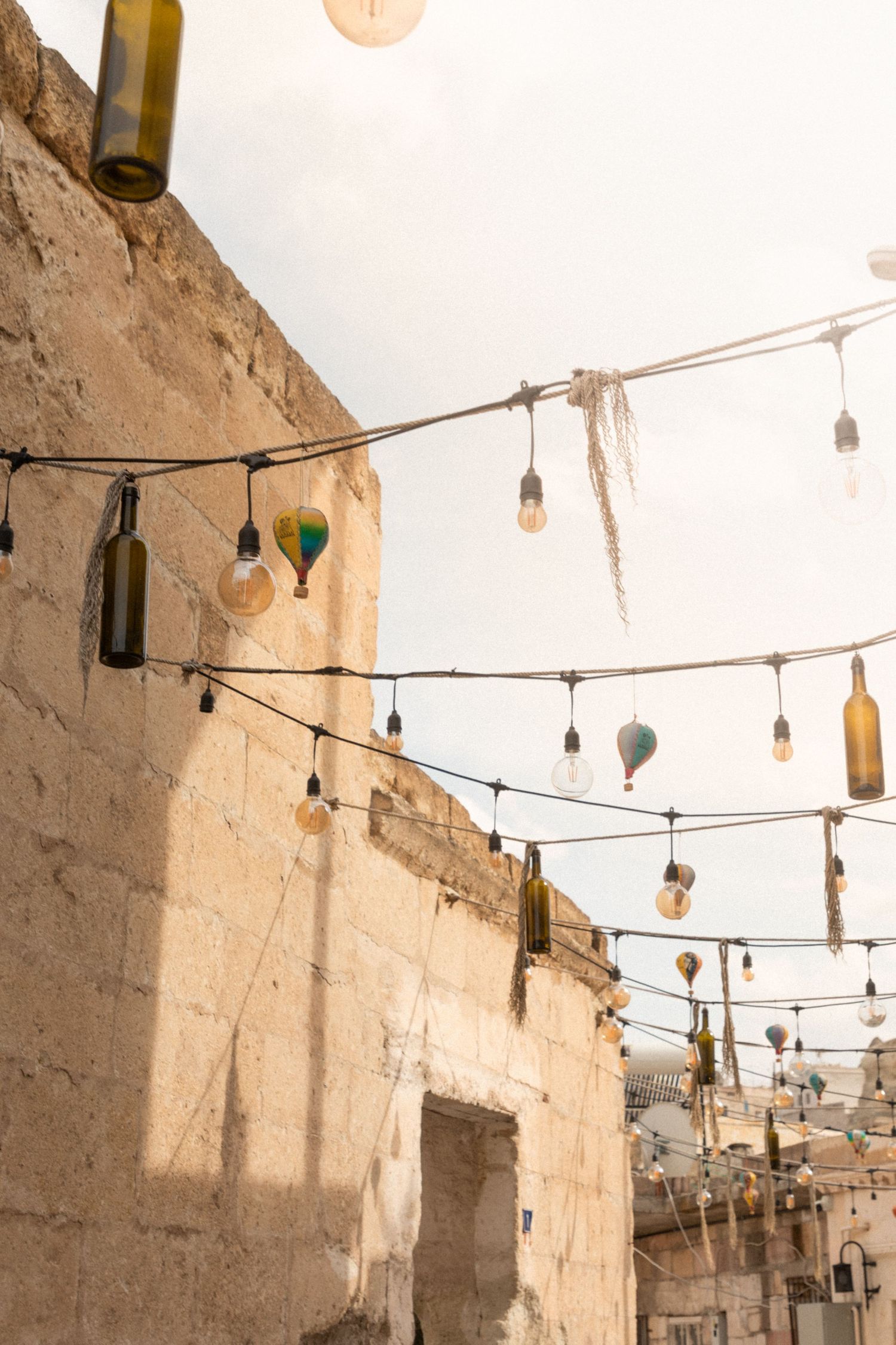 Decorative string lights with glass bottles and bulbs hanging between stone buildings in Cappadocia.