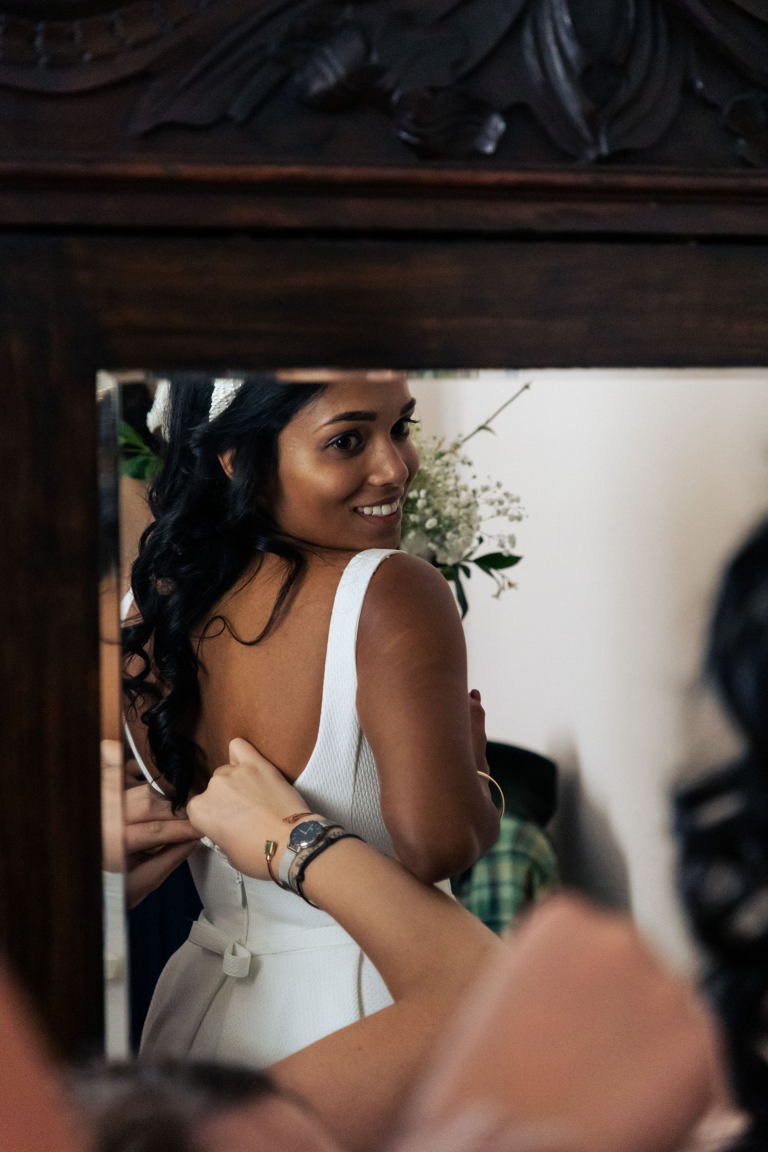 Smiling bride in white dress reflected in ornate wooden mirror while being helped by bridesmaid.