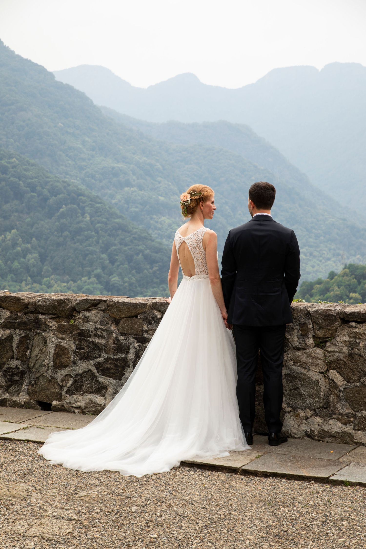 Bride and groom stand together at stone wall overlook with lush green mountain views on their wedding day.