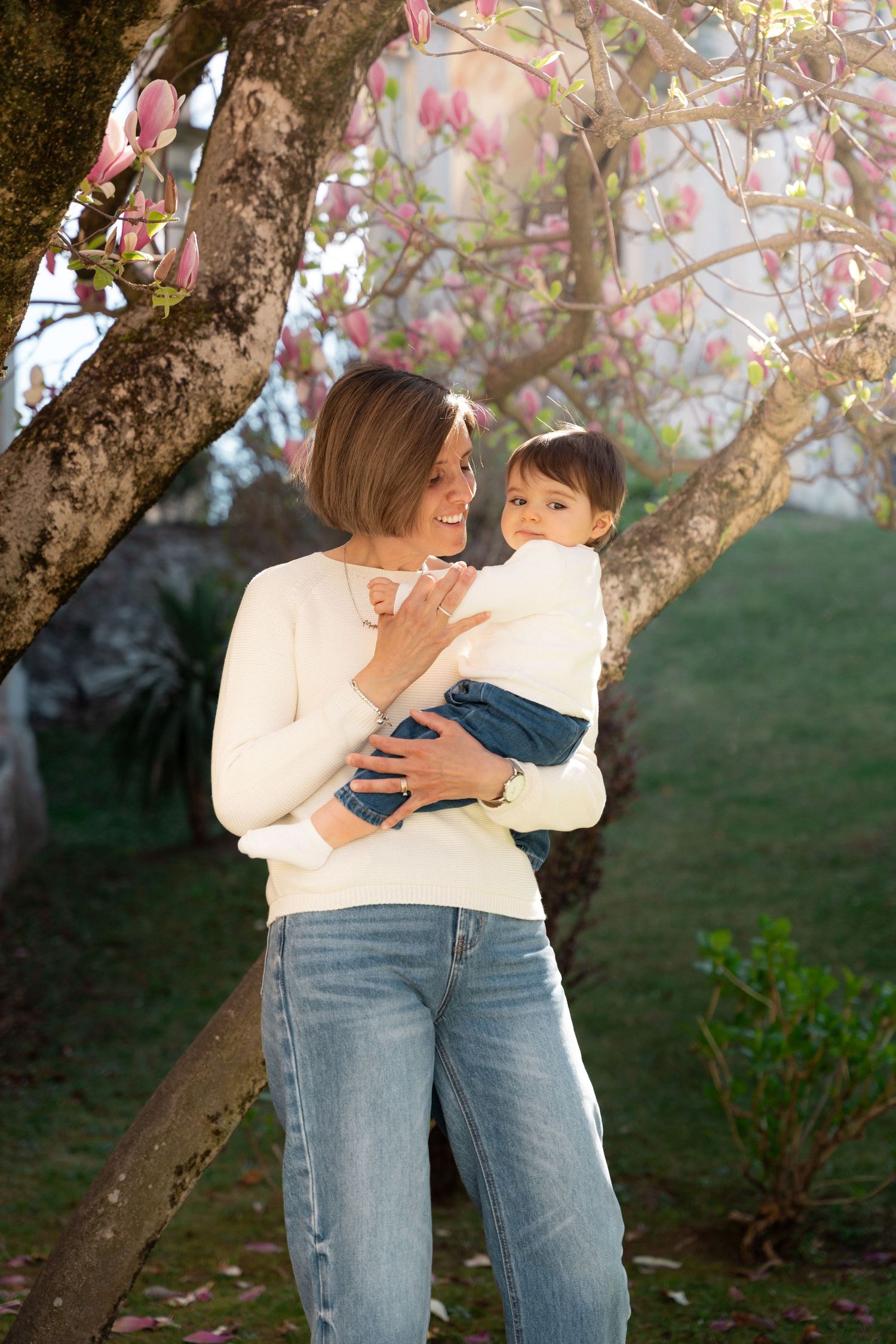 Mother holding baby boy under blooming pink magnolia tree in soft golden sunlight.