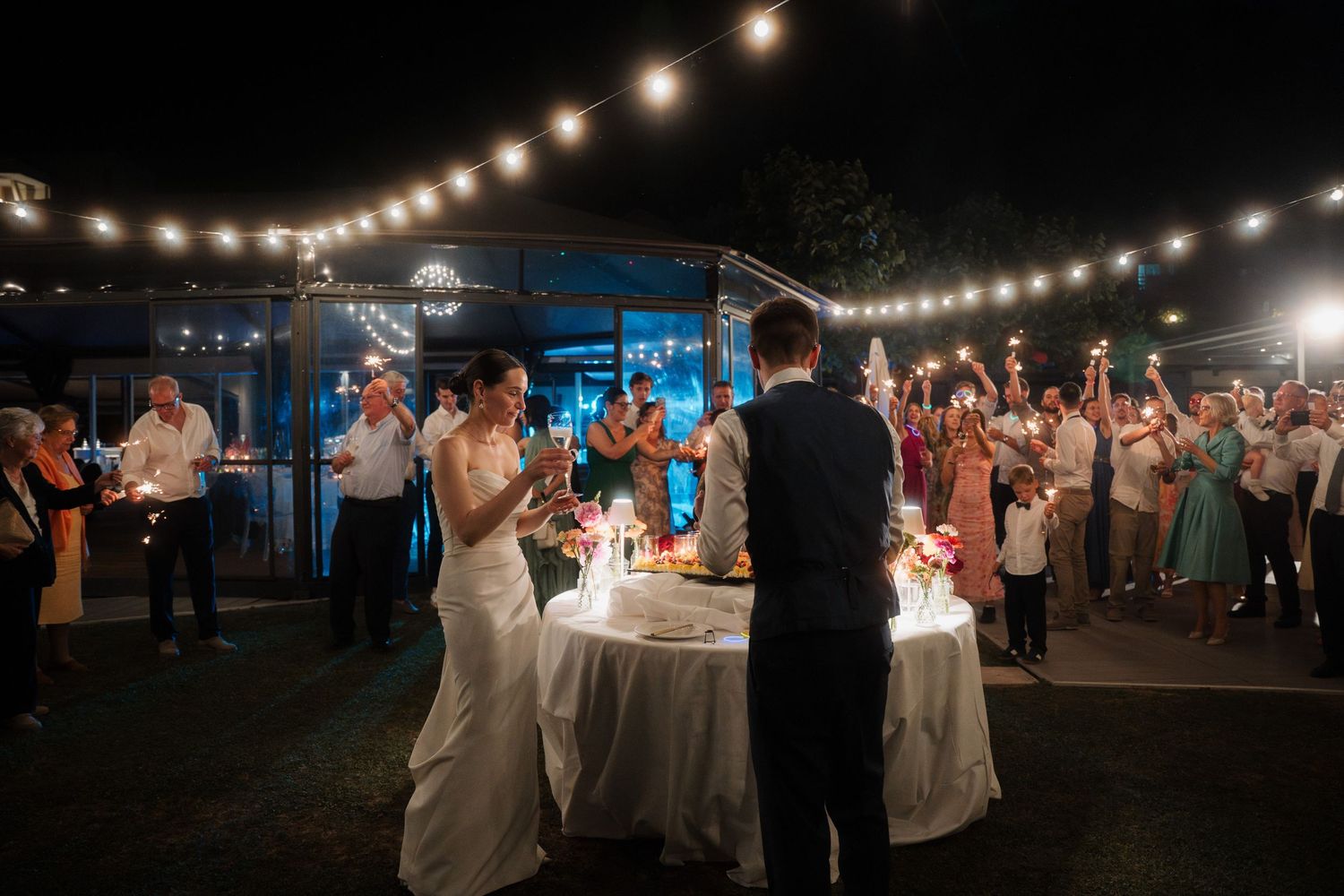 Bride and groom celebrate with sparklers surrounded by guests at an outdoor nighttime wedding reception.