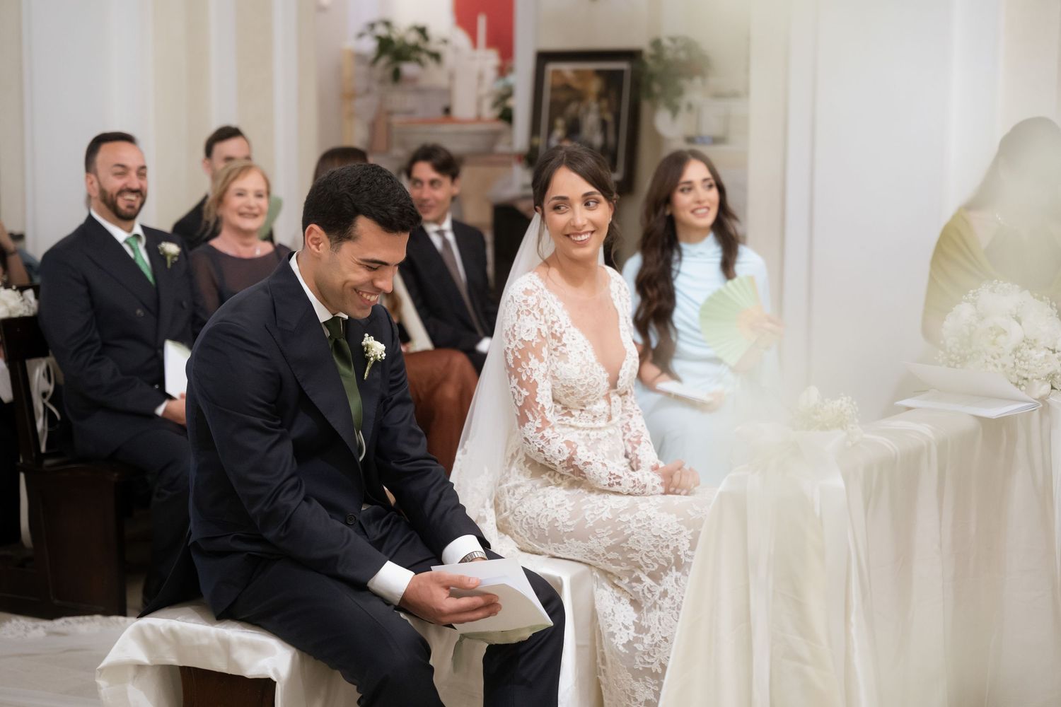 Bride and groom smiling during wedding ceremony seated at elegant white altar.
