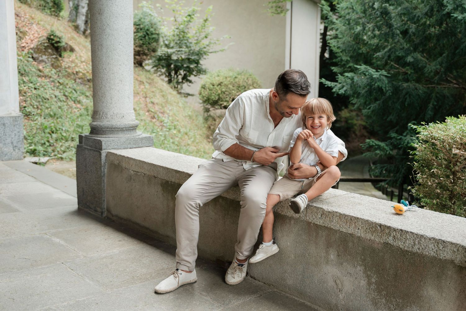Father and toddler sitting on stone ledge outdoors, sharing a tender moment in a garden setting.