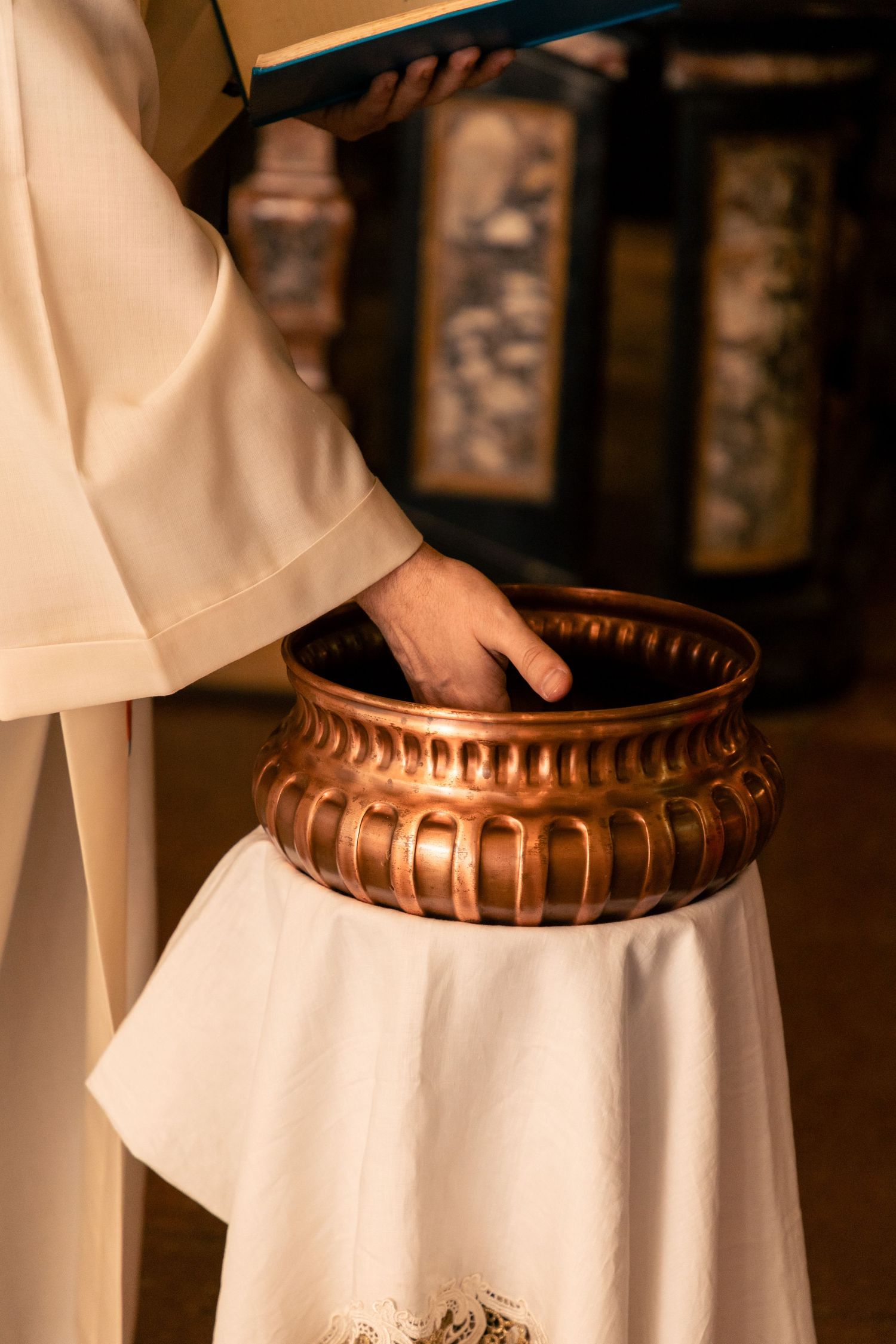 Priest in white robes holding ornate copper baptismal font during christening ceremony.