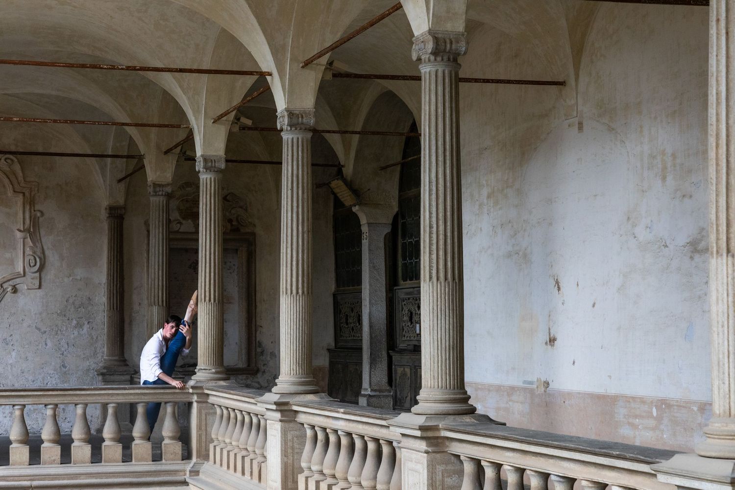 Dancer rests contemplatively on balustrade of weathered historic arcade with classical stone columns.