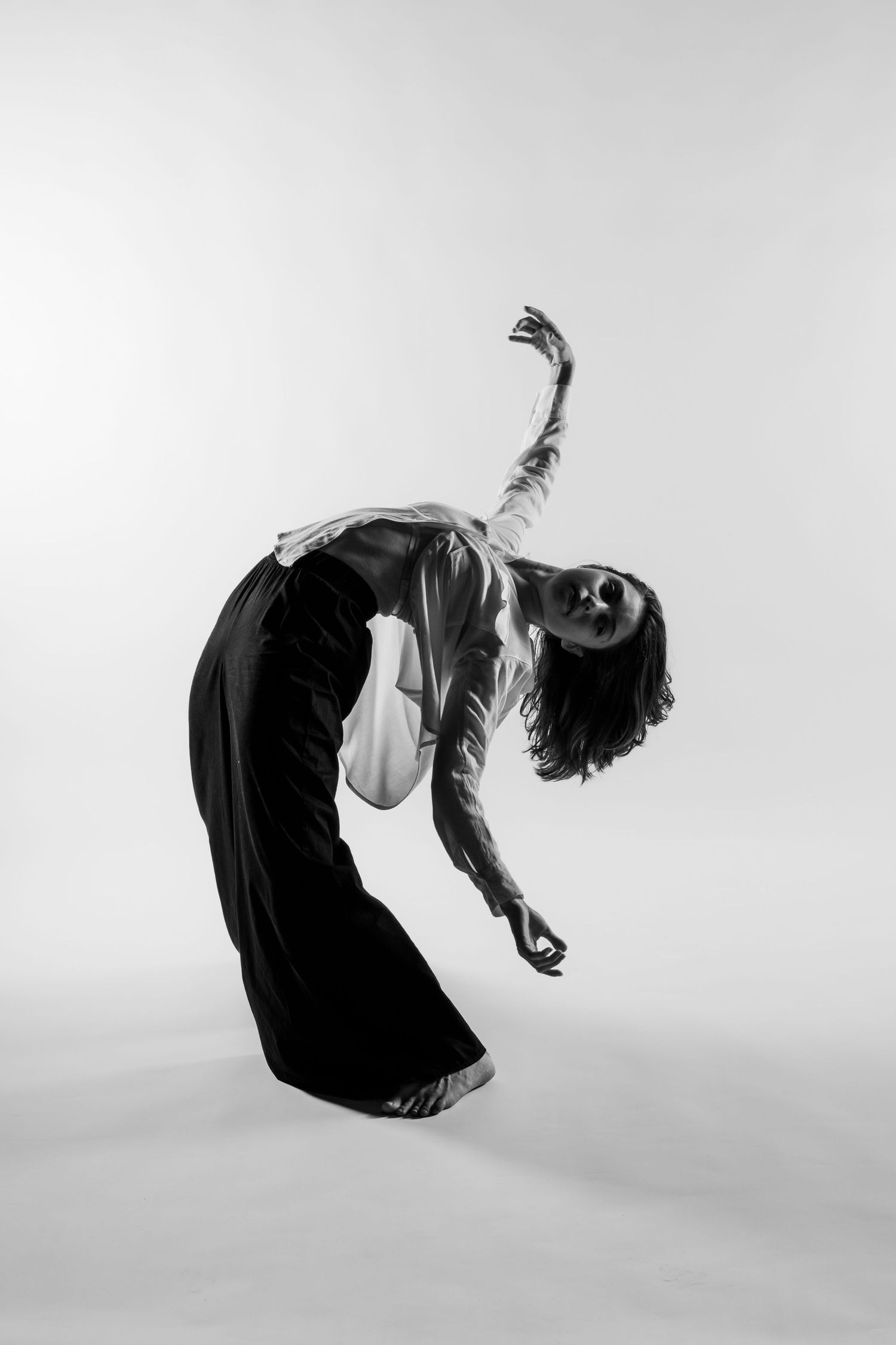 A dancer in black wide-leg pants and white top performs a deep backbend with one arm raised in a black and white photo.