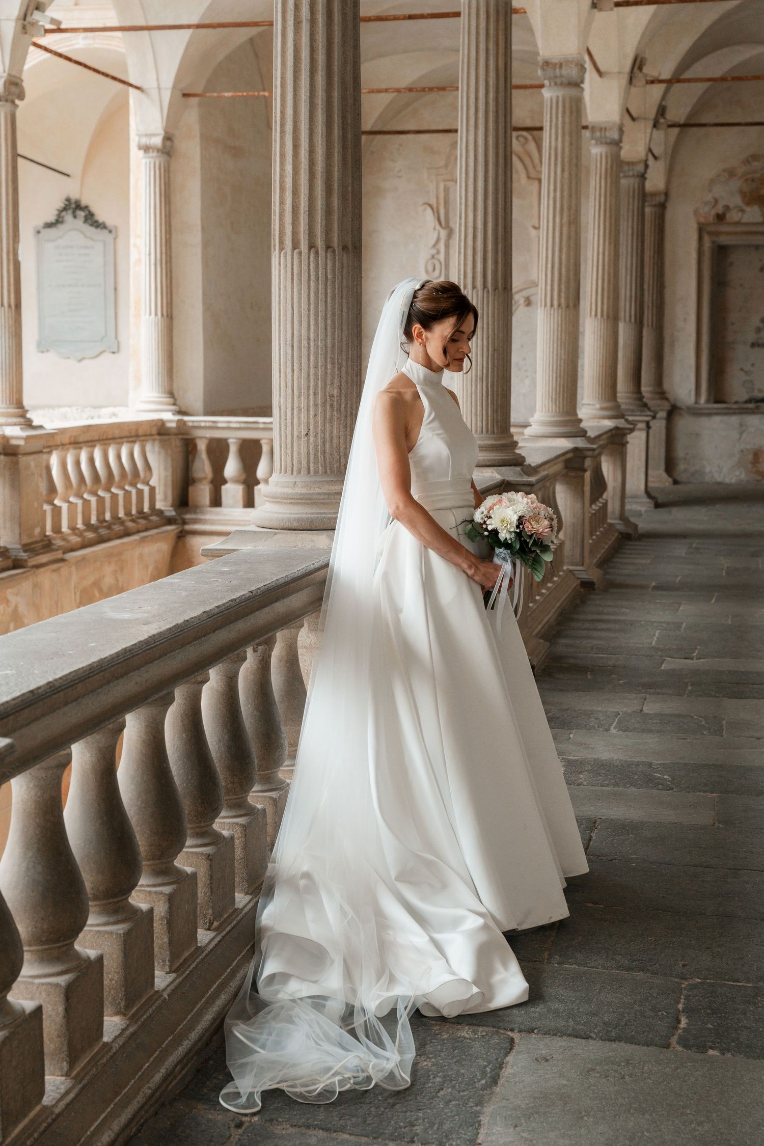 Bride in sleeveless white gown with veil poses gracefully on ornate stone staircase with classical columns.