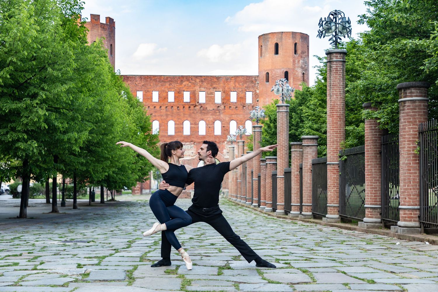 Ballet dance couple perform graceful arabesque pose on cobblestone path near ancient Roman Palatine Gates.