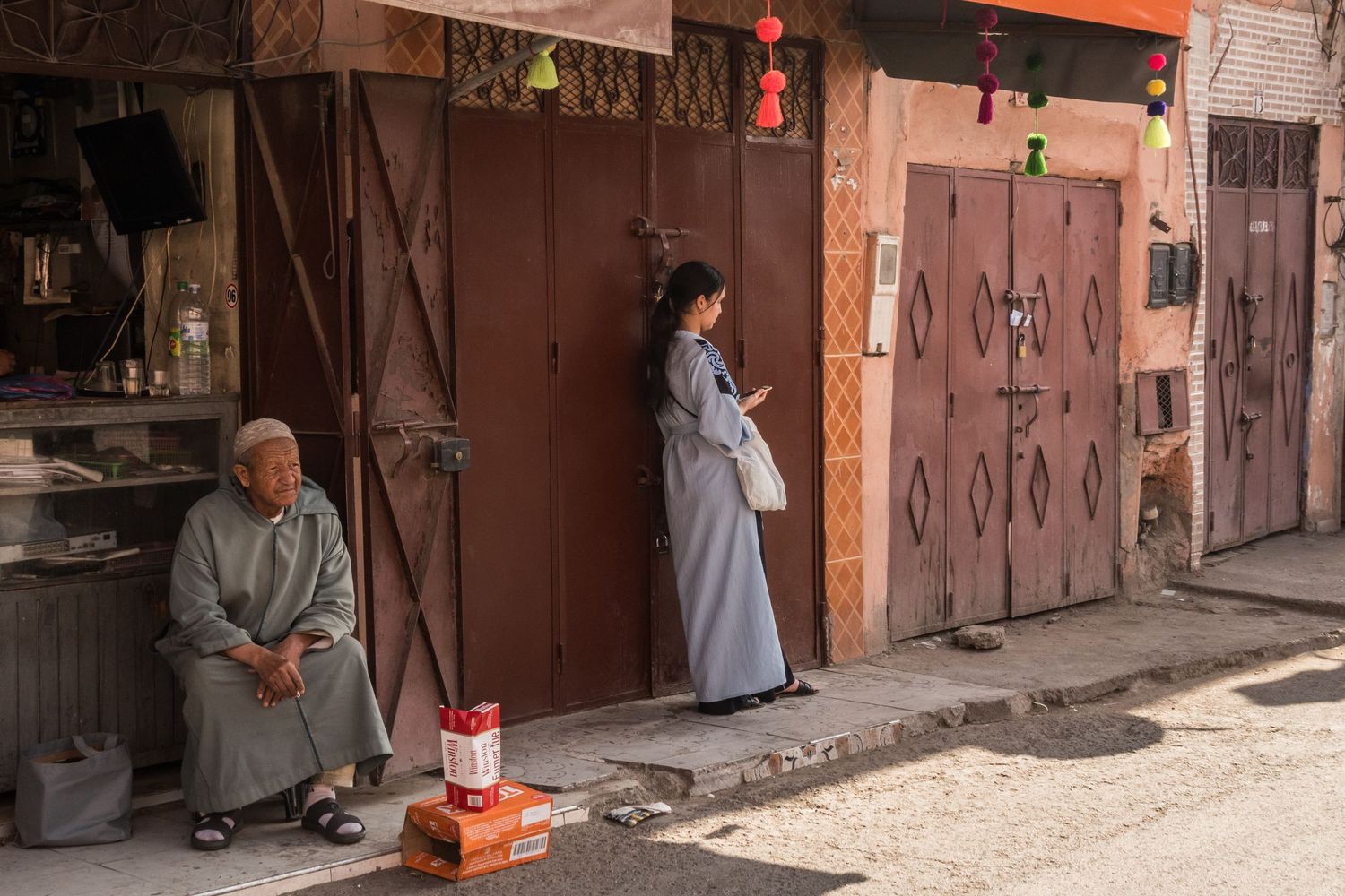 Two people in traditional Moroccan djellabas standing outside closed wooden doors on a street.