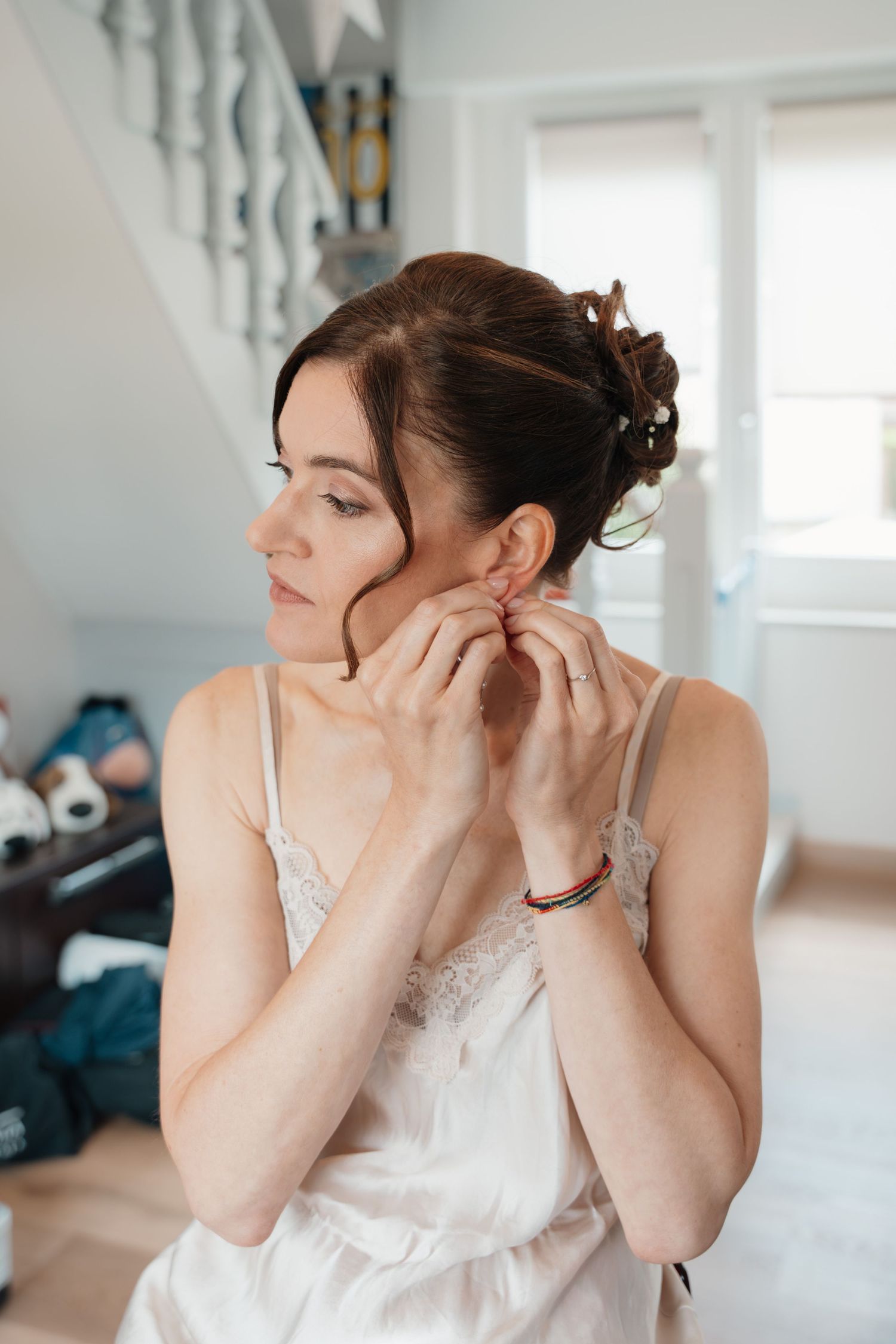 Beautiful bride in white slip dress puts on earrings while getting ready on her wedding morning.