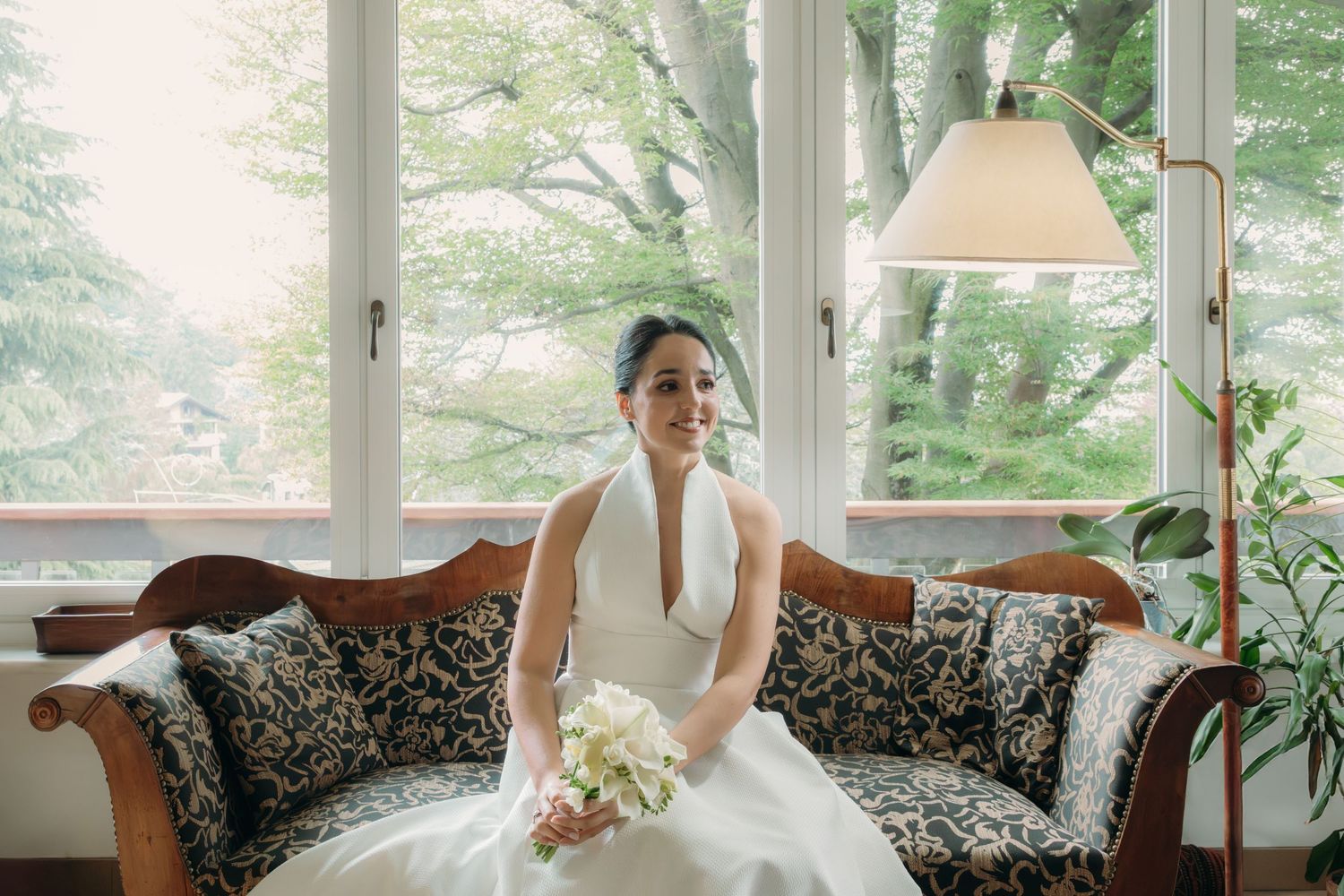 Smiling bride in sleek white halter gown sits on ornate sofa holding white bouquet by large windows.