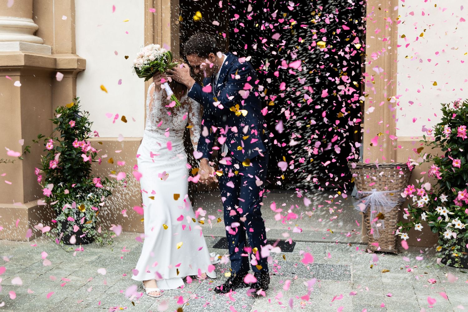 Bride and groom kiss in shower of pink confetti petals outside flower-adorned church entrance.