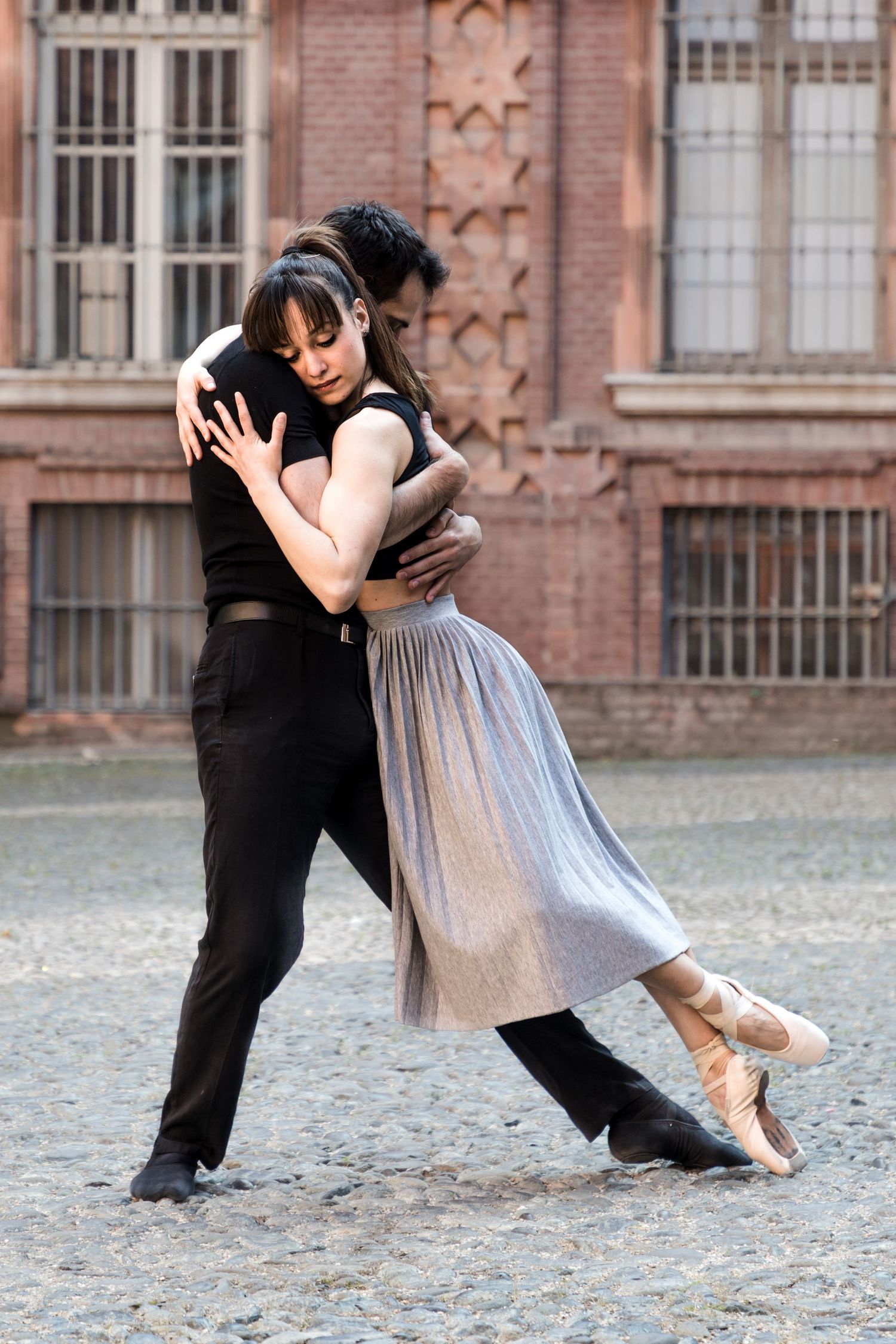 Male and female ballet dancers perform intimate tango pose on cobblestone street near historic building.