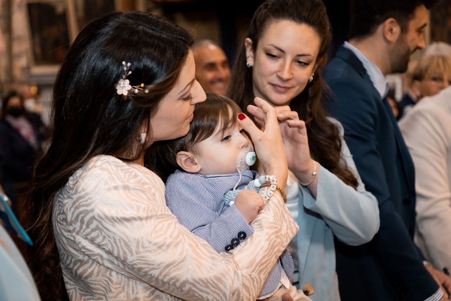Mother kissing her baby boy in a blue outfit while a woman assists during a baptism.