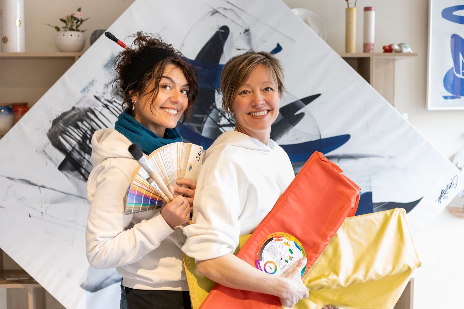 Two smiling women holding color swatches and art supplies in front of a large abstract painting in a studio.