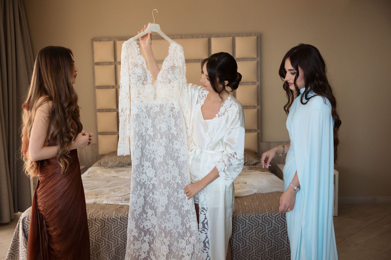Bride in white robe showing her lace wedding dress on hanger to two bridesmaids in a hotel room.