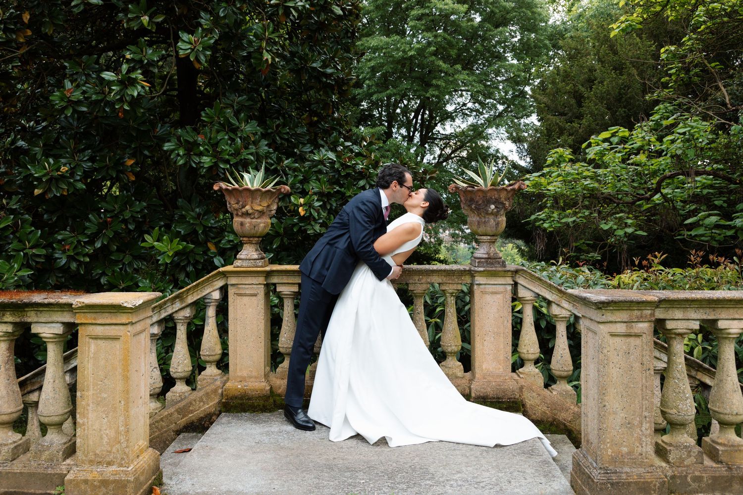 Bride and groom sharing passionate kiss on ornate stone balustrade in lush garden setting.