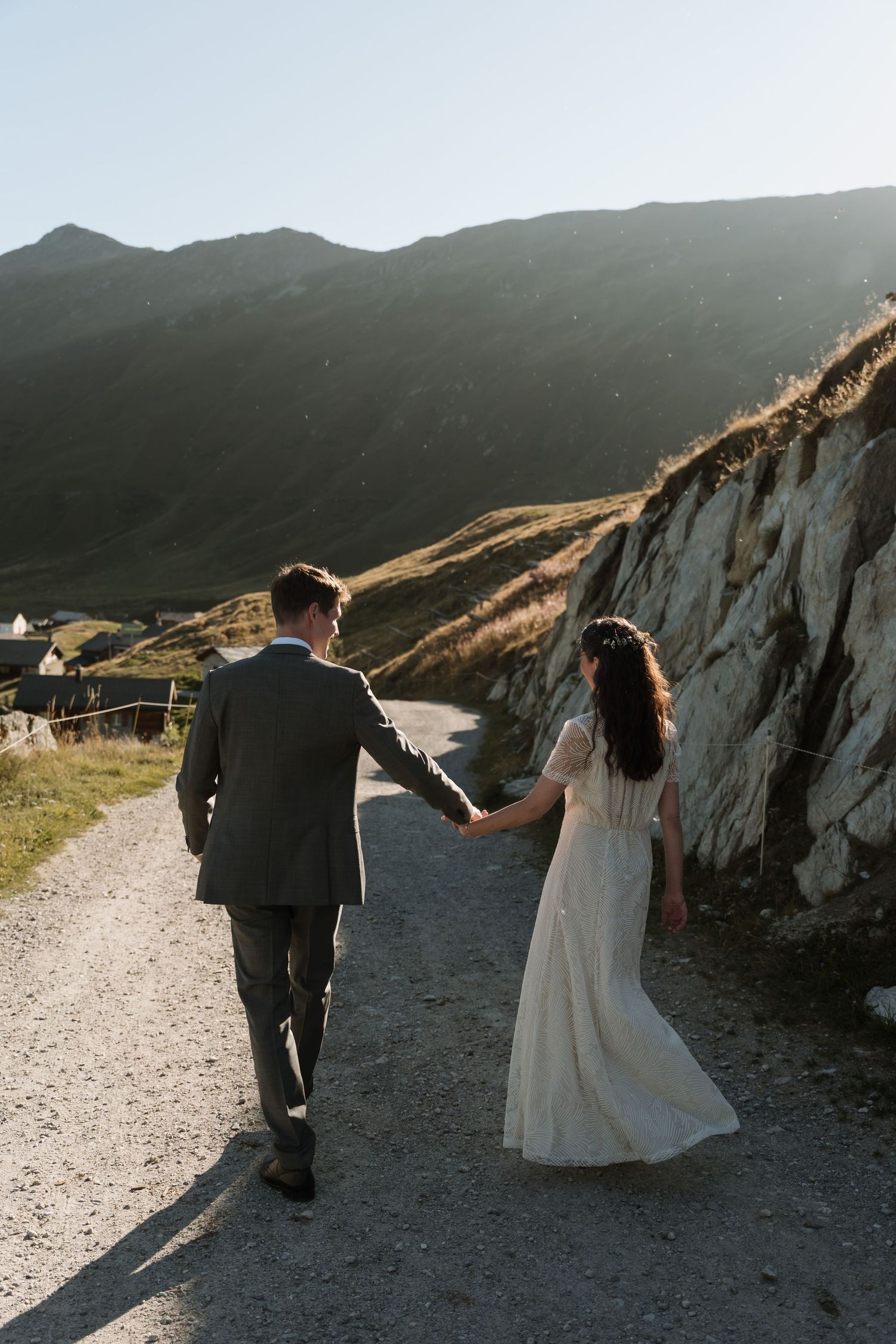 Bride and groom walking hand in hand on a gravel path with scenic mountain backdrop during golden hour.