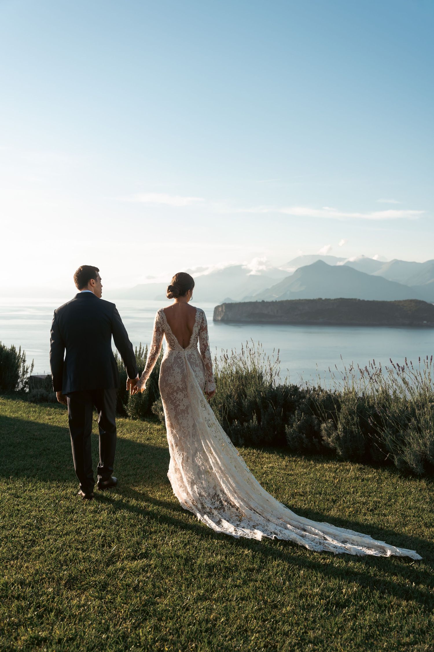 Bride and groom holding hands overlooking stunning coastal Mediterranean landscape at sunset.