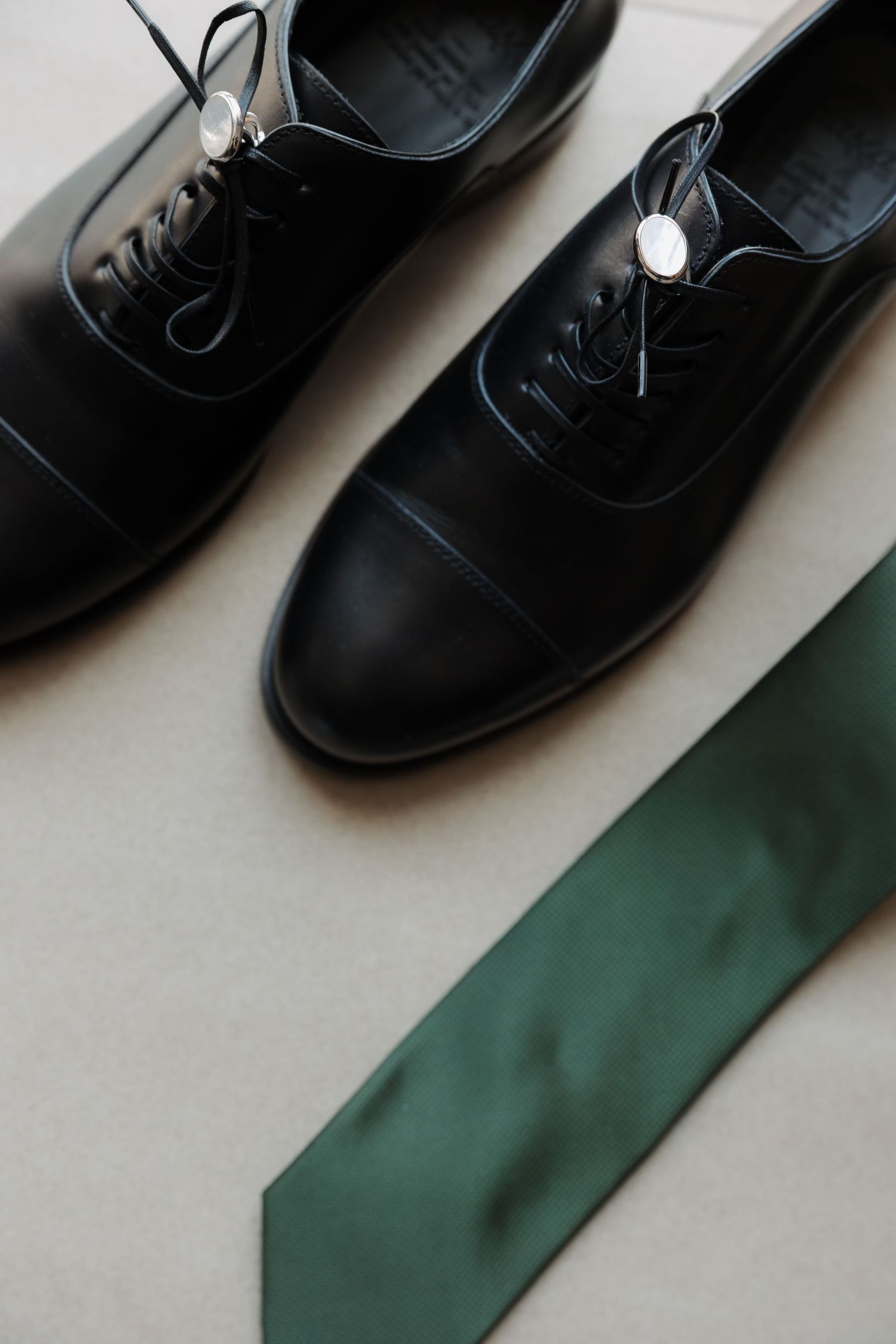 Black leather Oxford dress shoes with silver cufflinks and green silk tie arranged on beige surface.