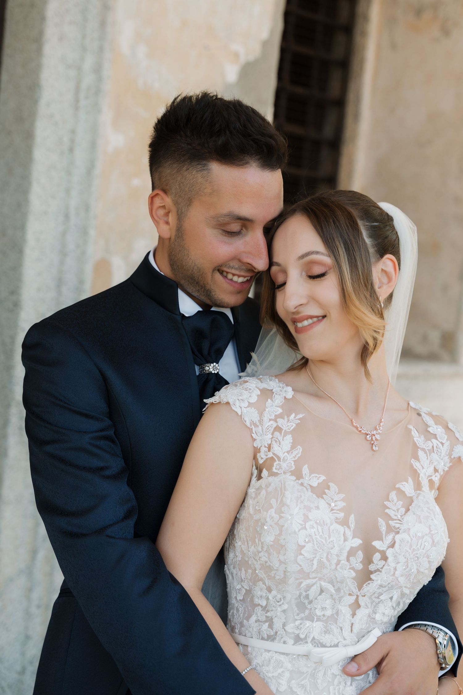 A smiling groom in a navy suit embraces his bride in a lace wedding dress and veil outdoors.