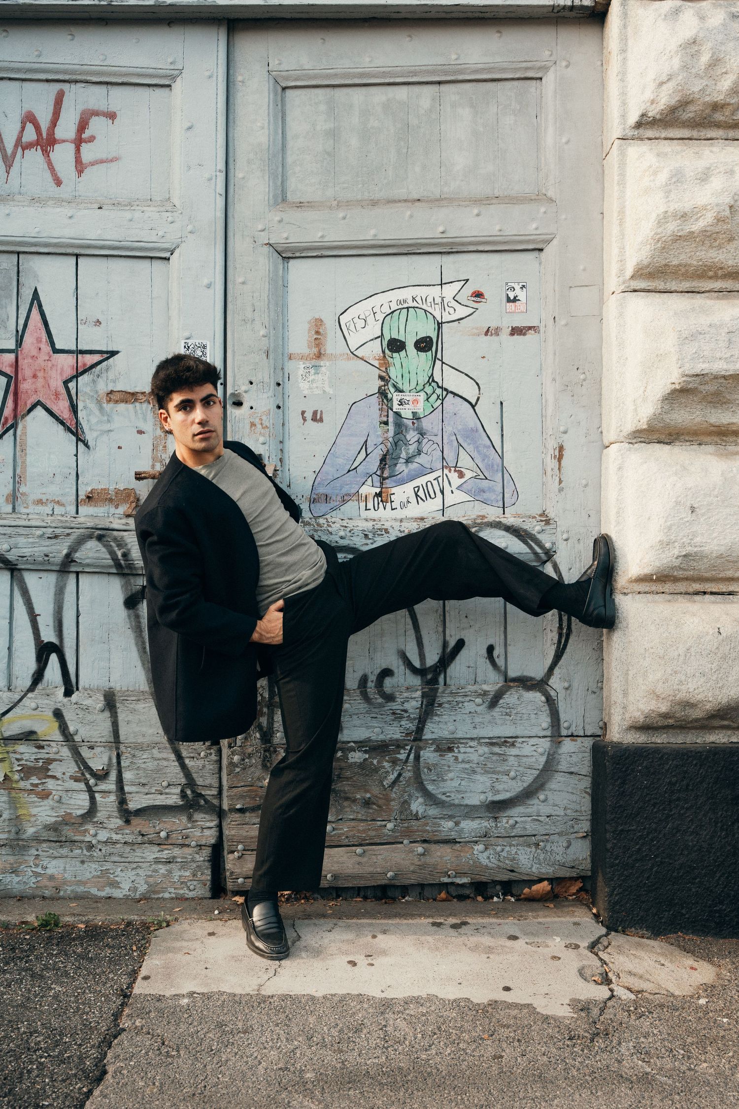 Young man in black coat posing with raised knee against graffiti-covered urban doorway.