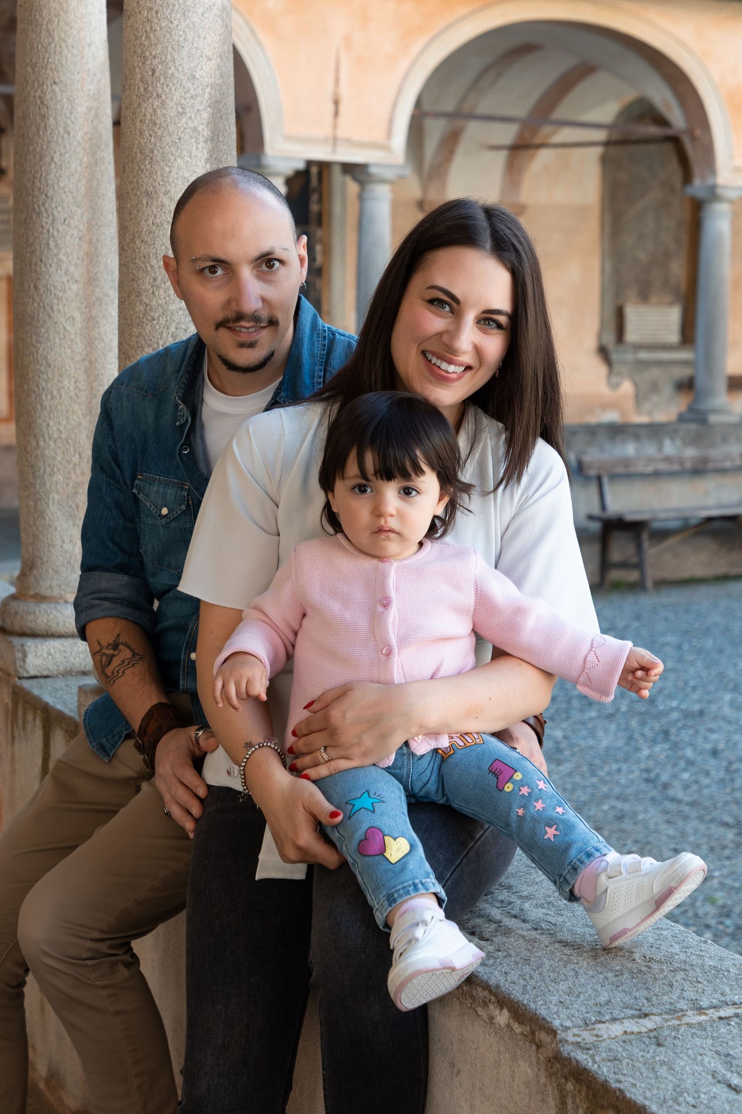 Happy family portrait with parents and toddler daughter sitting on stone ledge in historic Italian courtyard.