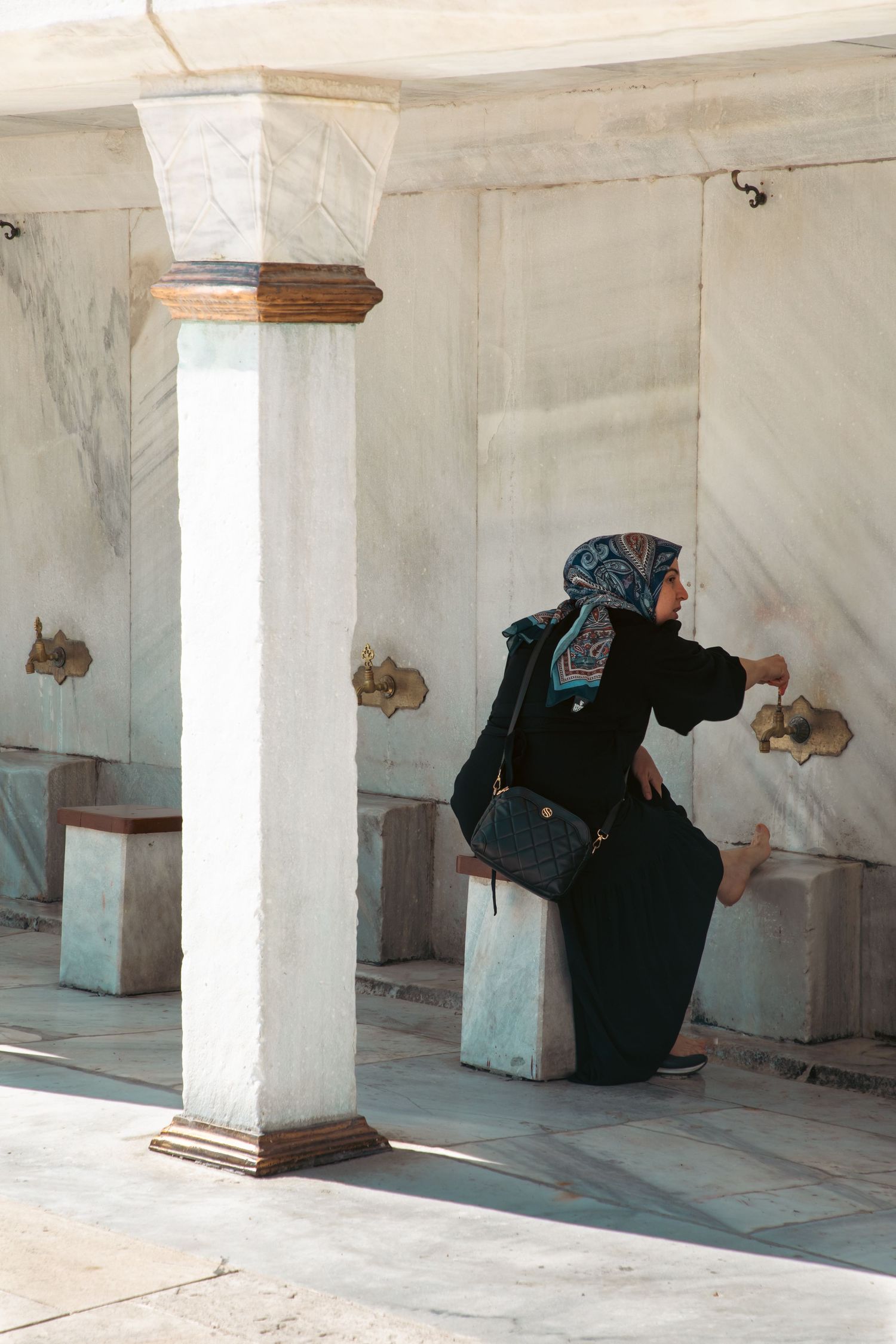 A woman in a hijab performing ritual ablution at a marble fountain outside a mosque in Istanbul.